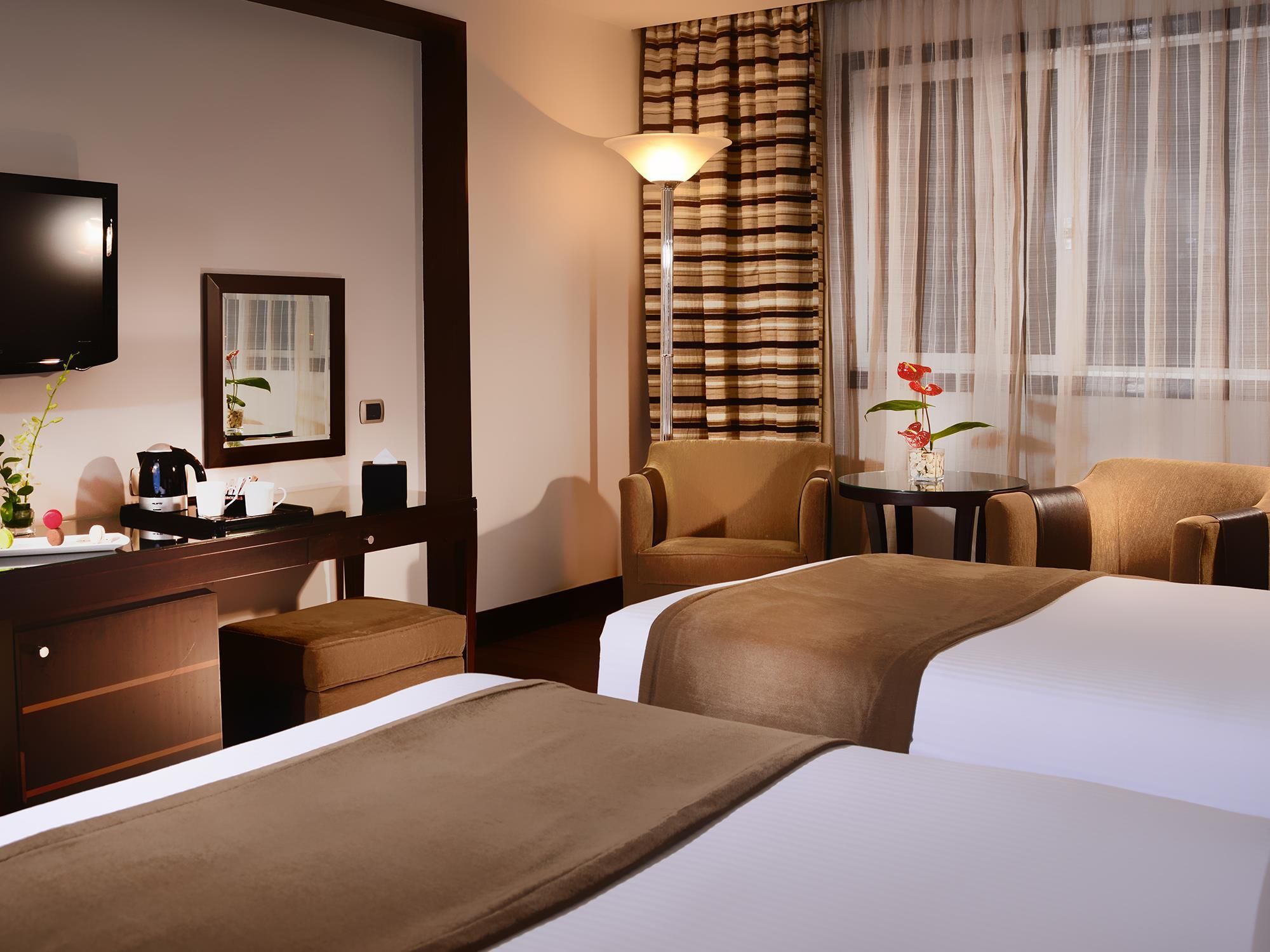 Habitación de hotel con dos camas, un escritorio con cafetera, una silla, televisor y ventana con cortinas.