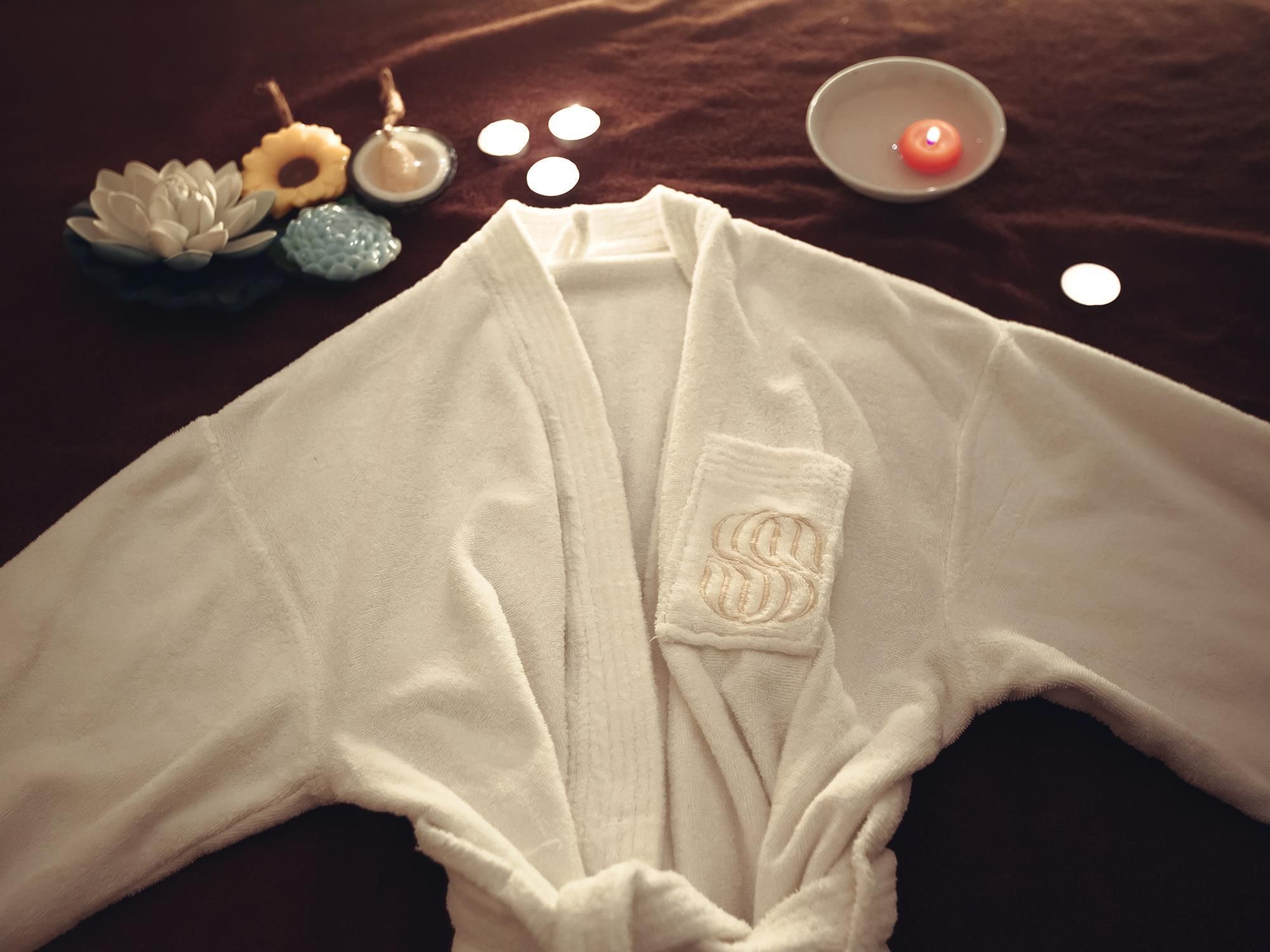 A white bathrobe with a pocket on the front, placed on a dark surface with a lit candle and decorative items nearby.