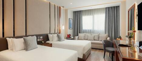 A hotel room with two beds, a sofa, a desk, and a large window with curtains.