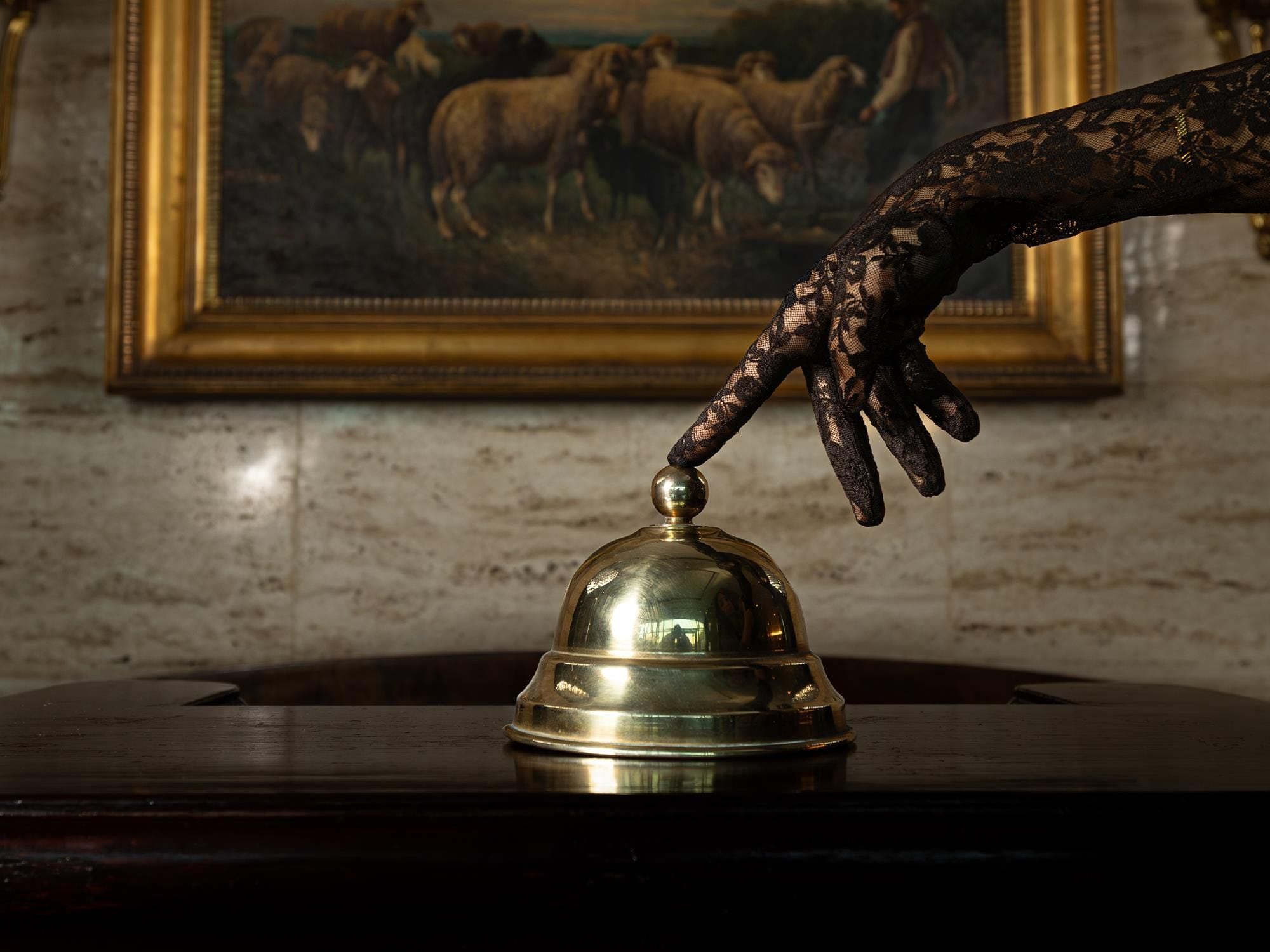 A hand reaching towards a bell on a wooden surface, with a framed painting in the background.