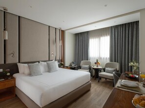 Habitación de hotel con una cama grande, un escritorio con una silla, una mesita con un frutero y una ventana con cortinas.
