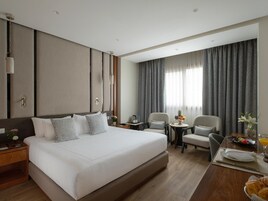 Habitación de hotel con una cama grande, un escritorio con una silla, una mesita con un frutero y una ventana con cortinas.