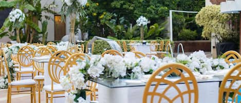 An outdoor dining area with round tables and chairs, surrounded by greenery and flowers.