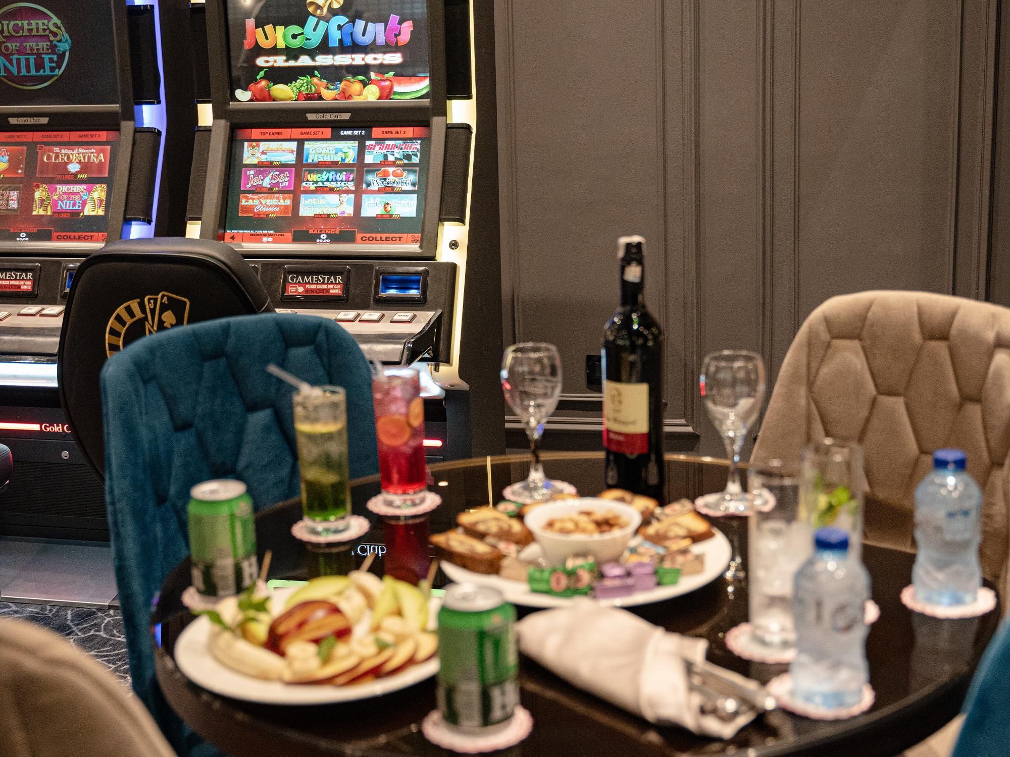 A dining table set with food, drinks, and a bottle of wine, with slot machines in the background.