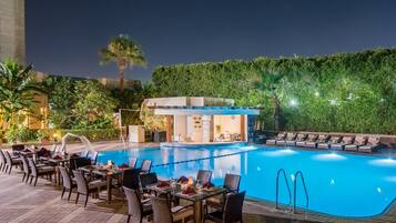 A poolside area with a dining setup and a small building, surrounded by greenery.