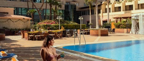 Un área junto a la piscina con sillones de descanso, una piscina y un edificio de varios pisos al fondo.