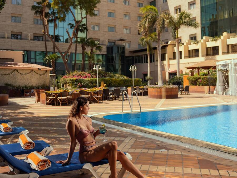 Un área junto a la piscina con sillones de descanso, una piscina y un edificio de varios pisos al fondo.