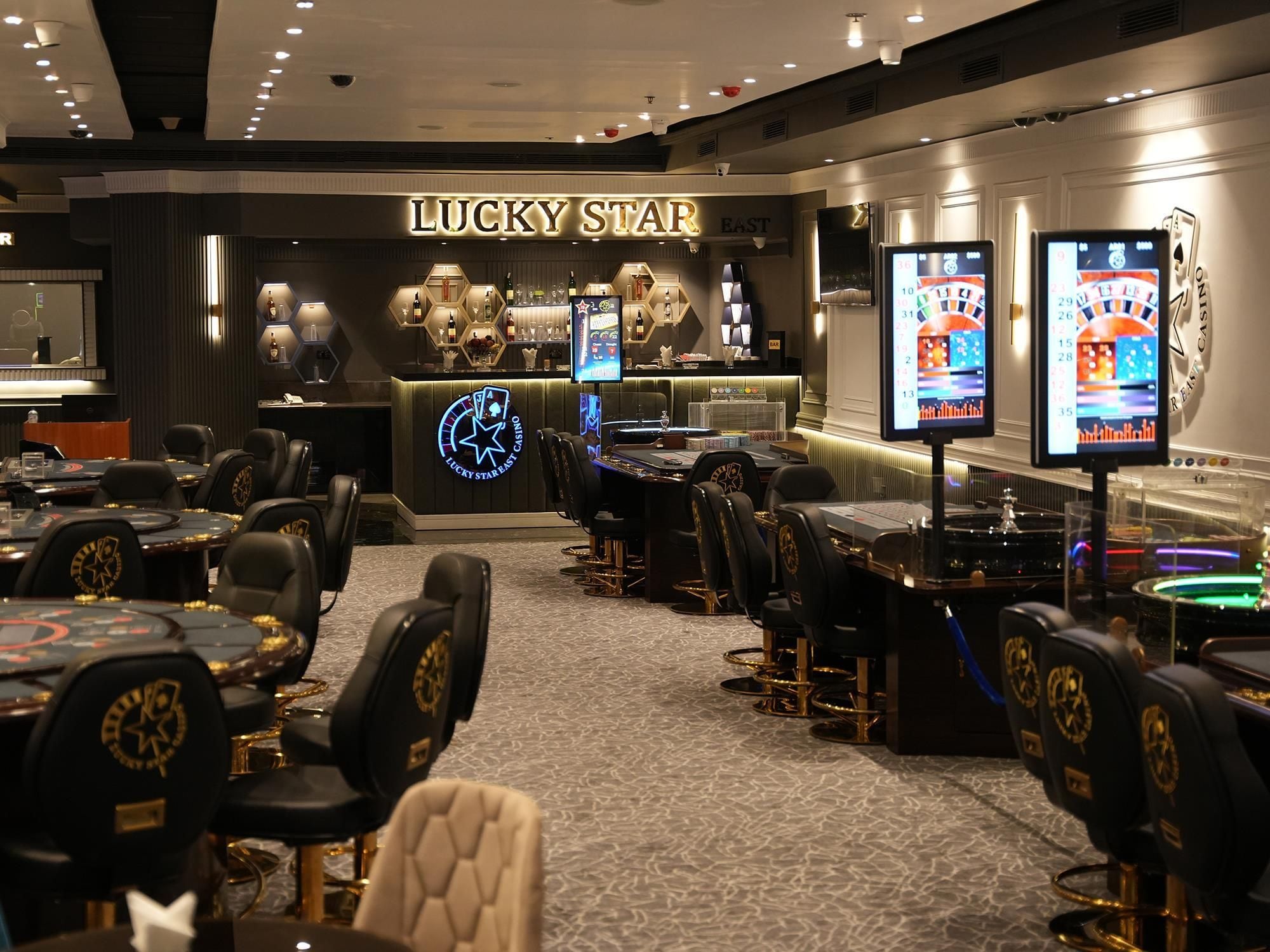 Zona de casino con varias mesas de juego y sillas, un bar y máquinas tragamonedas.
