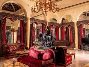 Un interior lujoso con una gran araña, columnas ornamentales y sofás rojos y cómodos.
