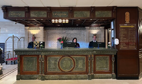 Una recepción con encimera de mármol, dos recepcionistas, un reloj y un cartel que enumera los servicios del hotel.