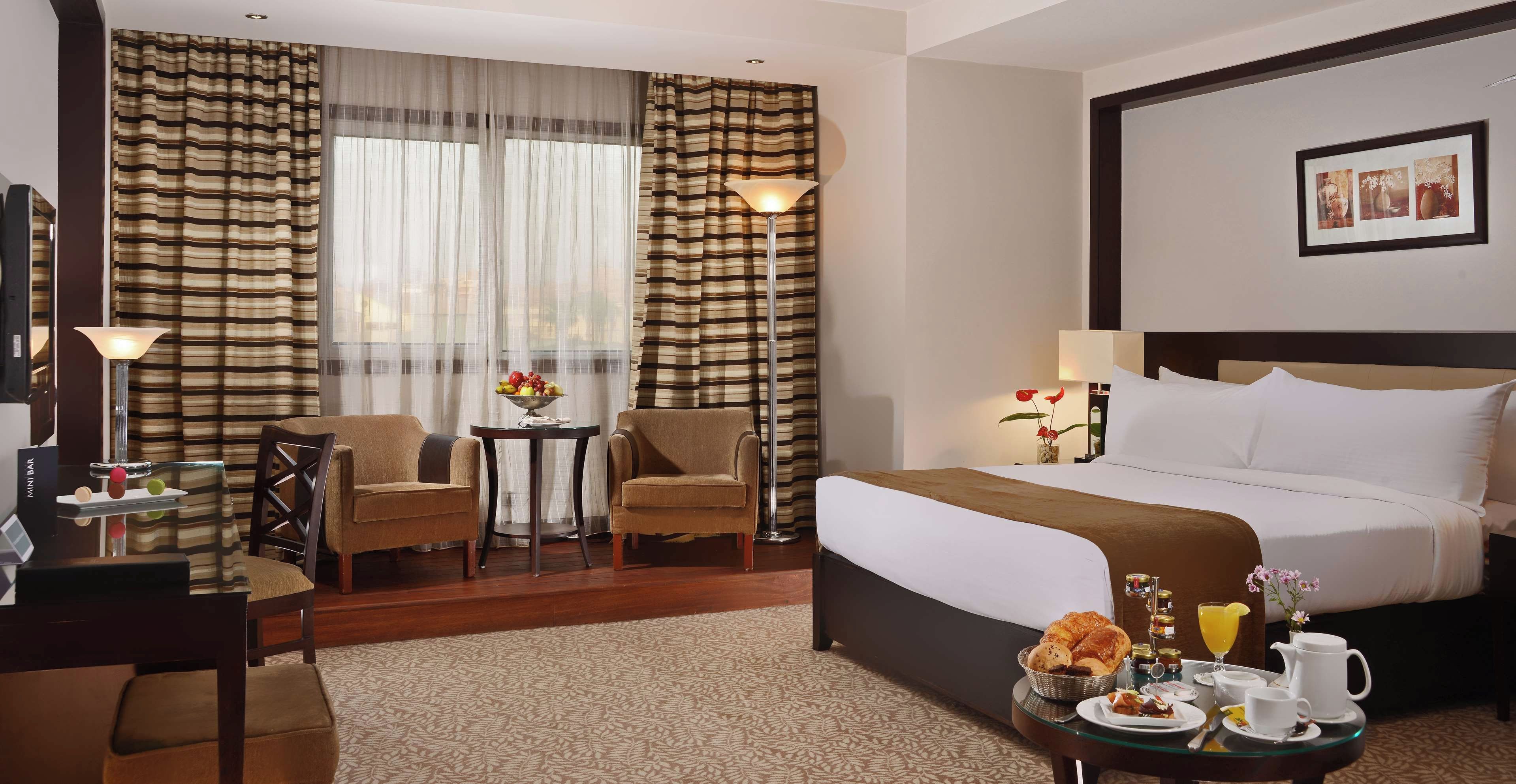 A hotel room with a bed, two armchairs, a small table with a fruit bowl, a lamp, and a framed picture on the wall.