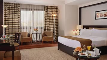A hotel room with a bed, two armchairs, a small table with a fruit bowl, a lamp, and a framed picture on the wall.