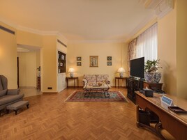 Ein Wohnzimmer mit einer Couch, einem Couchtisch und einem Fernseher auf einem Schrank. Es gibt einen Schreibtisch mit einem Computer und einem Telefon. Der Raum hat Holzböden und einen Teppich mit Muster.