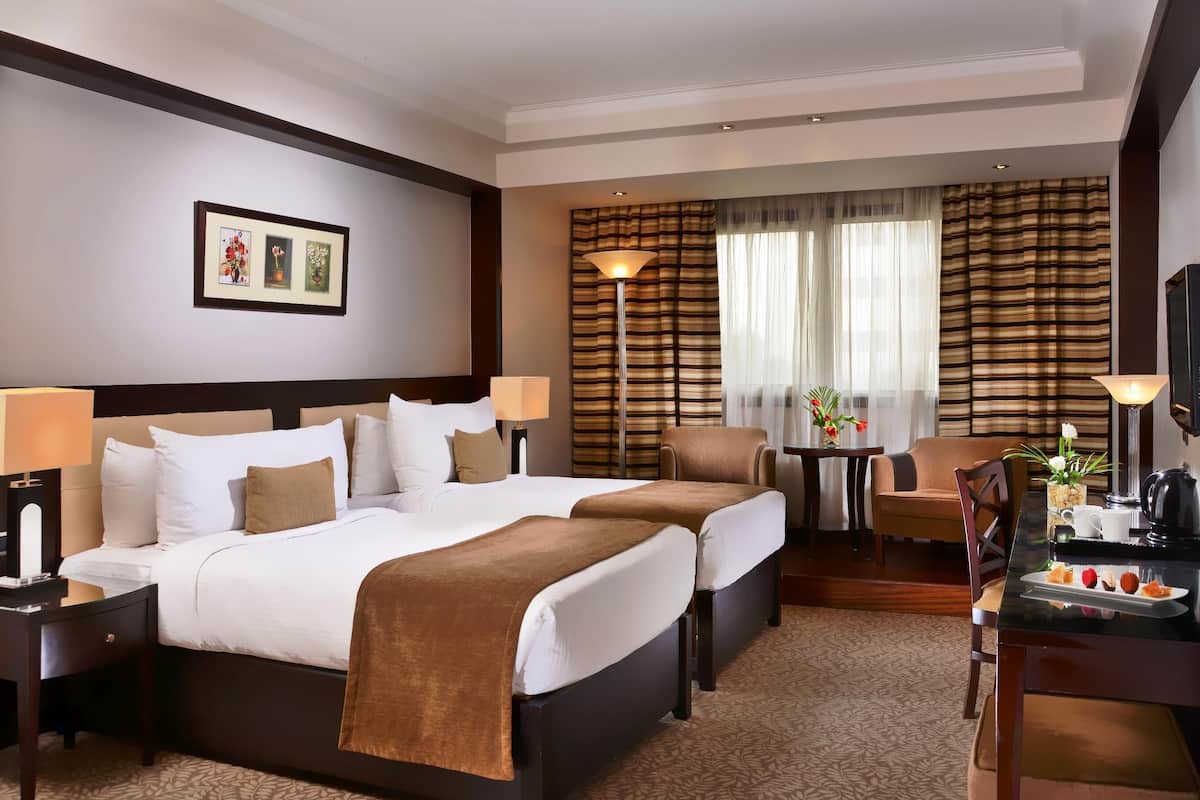 Habitación de hotel con una cama grande, dos mesitas de noche con lámparas, una pequeña mesa con un jarrón de flores, una silla y un escritorio con una cafetera y una bandeja de fruta.