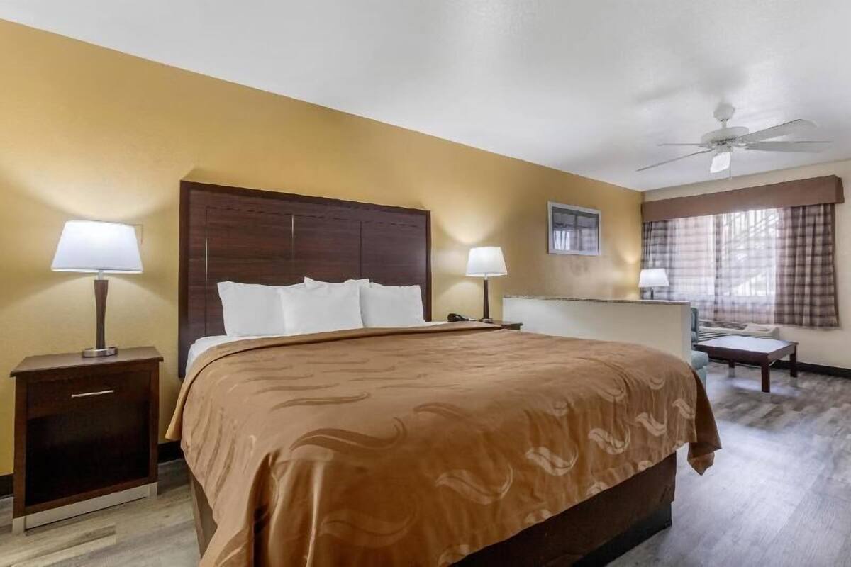 A hotel room with a large bed, two bedside tables with lamps, a ceiling fan, a window with curtains, and a small table.