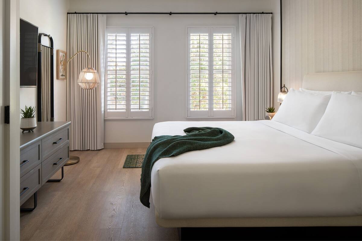 A neatly arranged bedroom with a bed, a dresser, a lamp, and two windows with curtains.