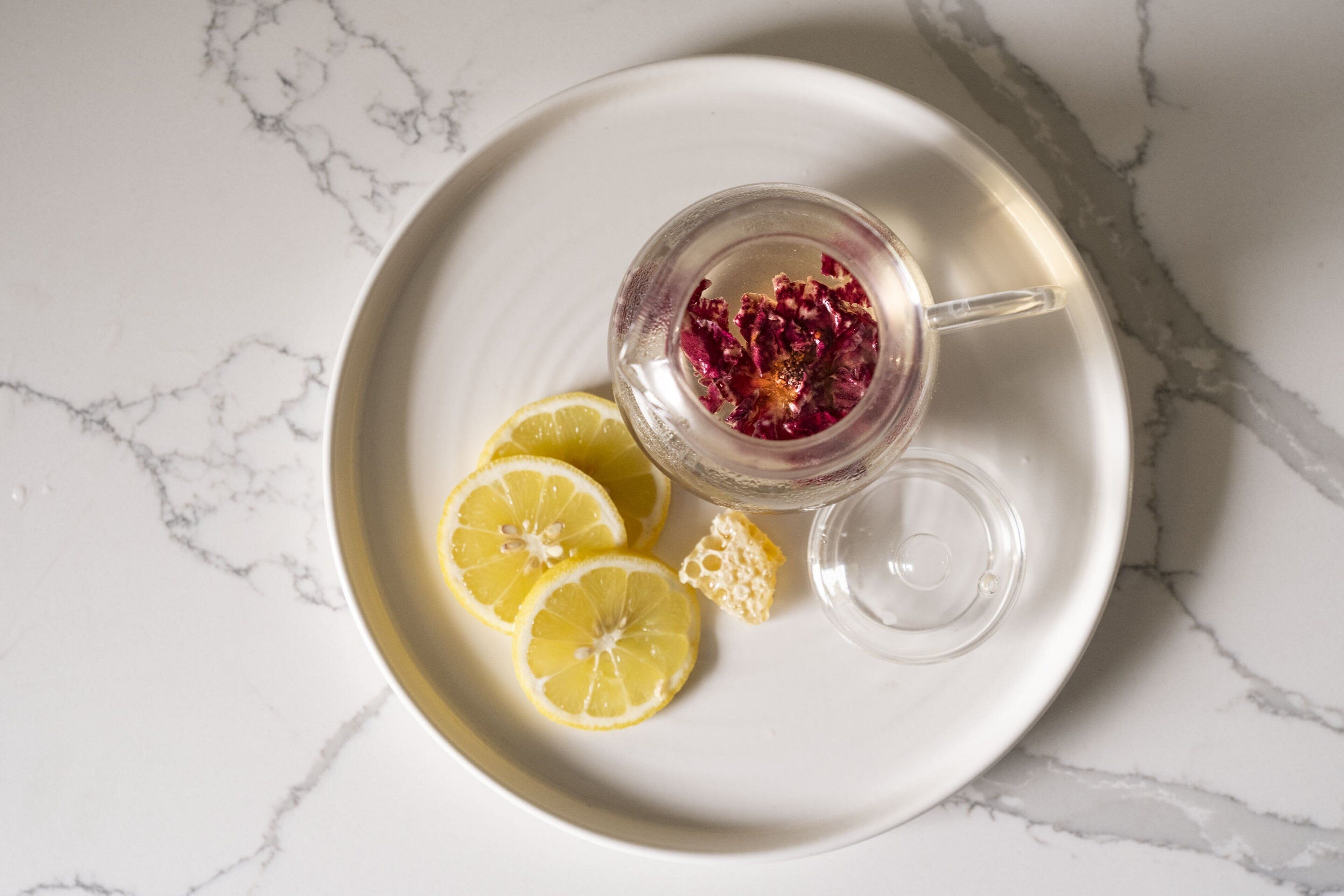 A clear glass mug with dried flowers, lemon slices, and a small glass on a white plate with a marble pattern background.