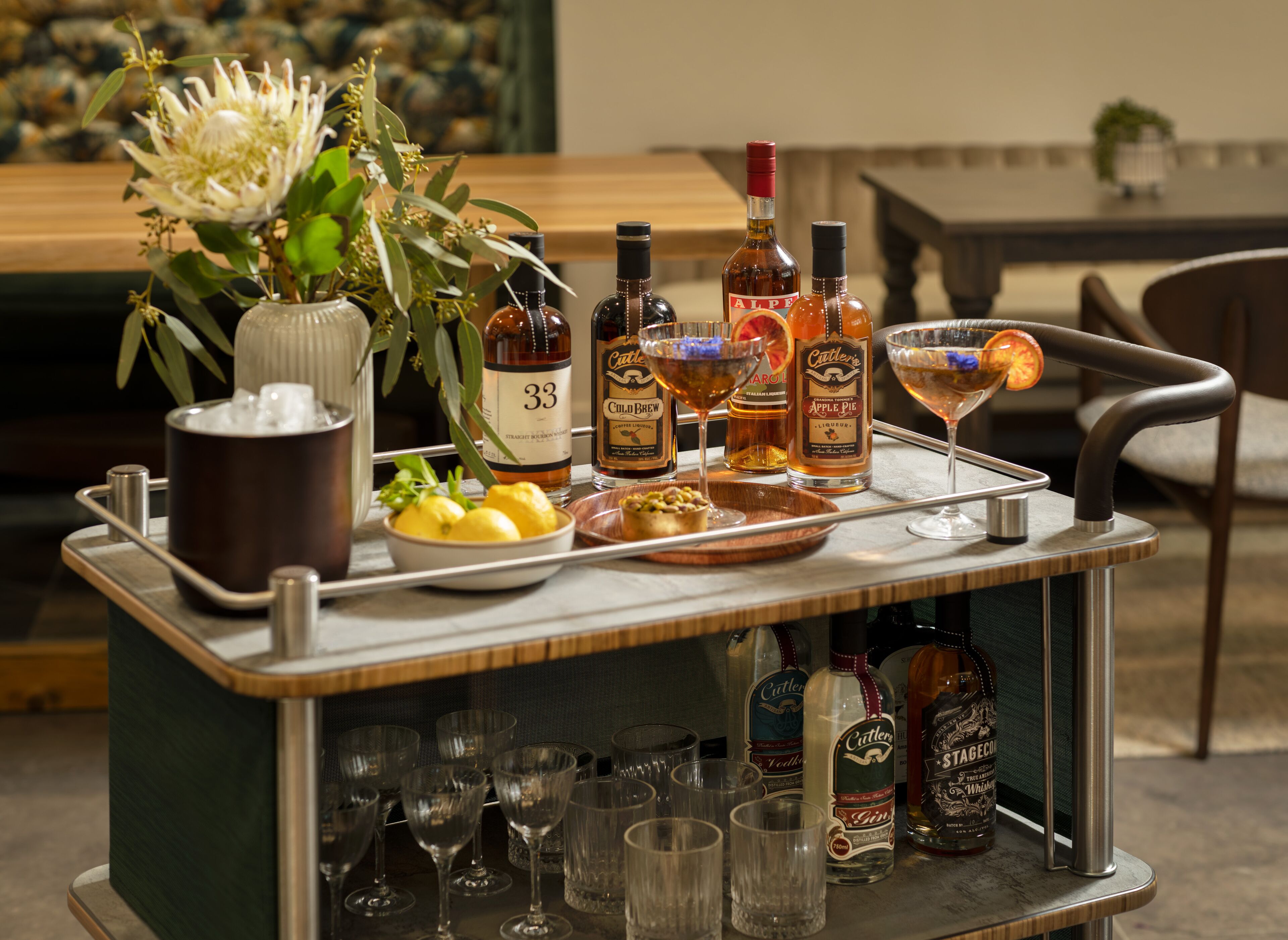 A bar cart with various bottles and glasses, a bowl of lemons, and a vase with flowers.