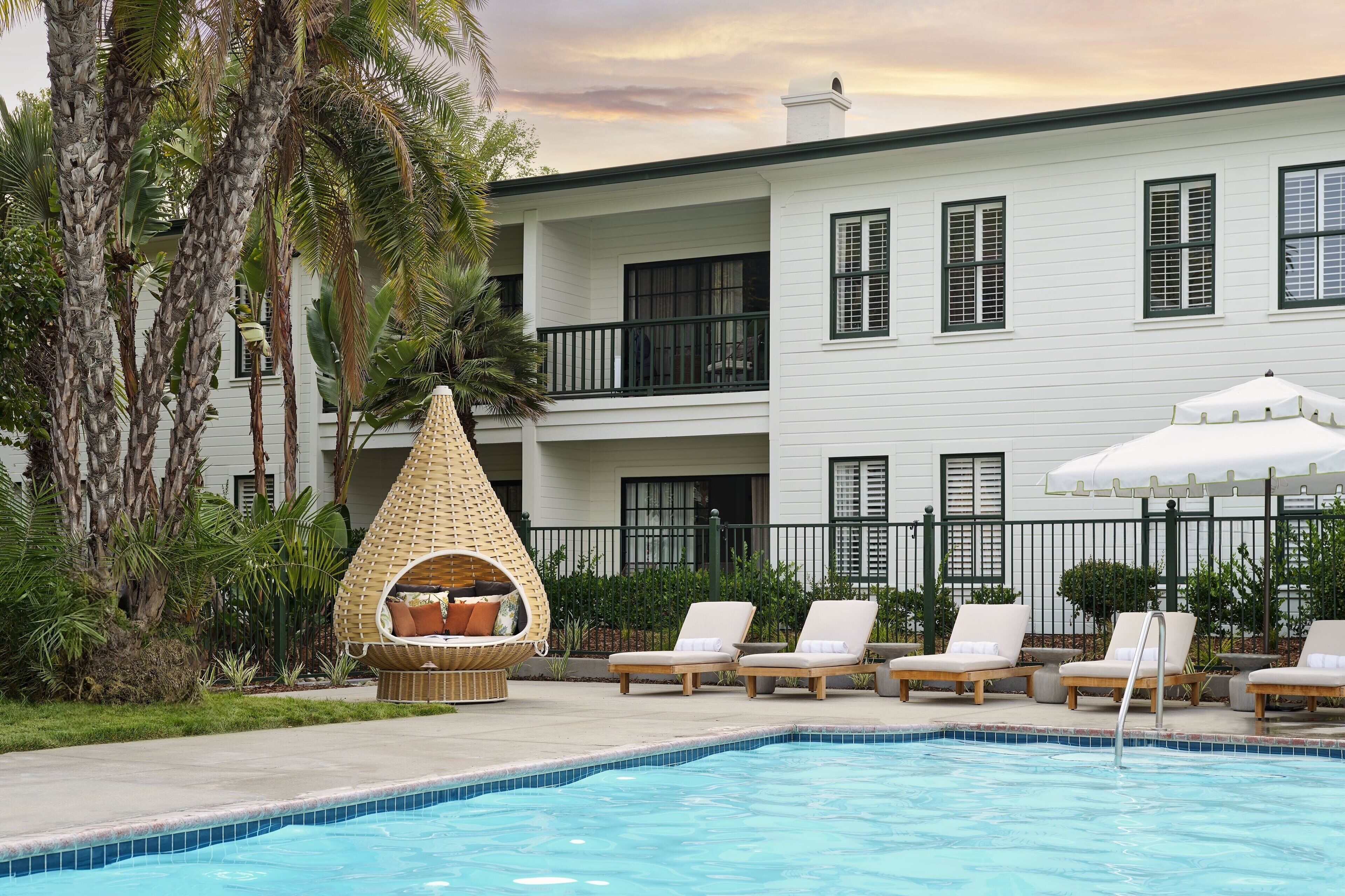 A pool area with lounge chairs, a wicker chair, and a white building with shutters.