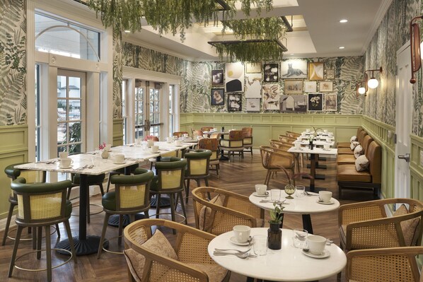 Um restaurante com várias mesas e cadeiras, uma parede verde com quadros e plantas penduradas.