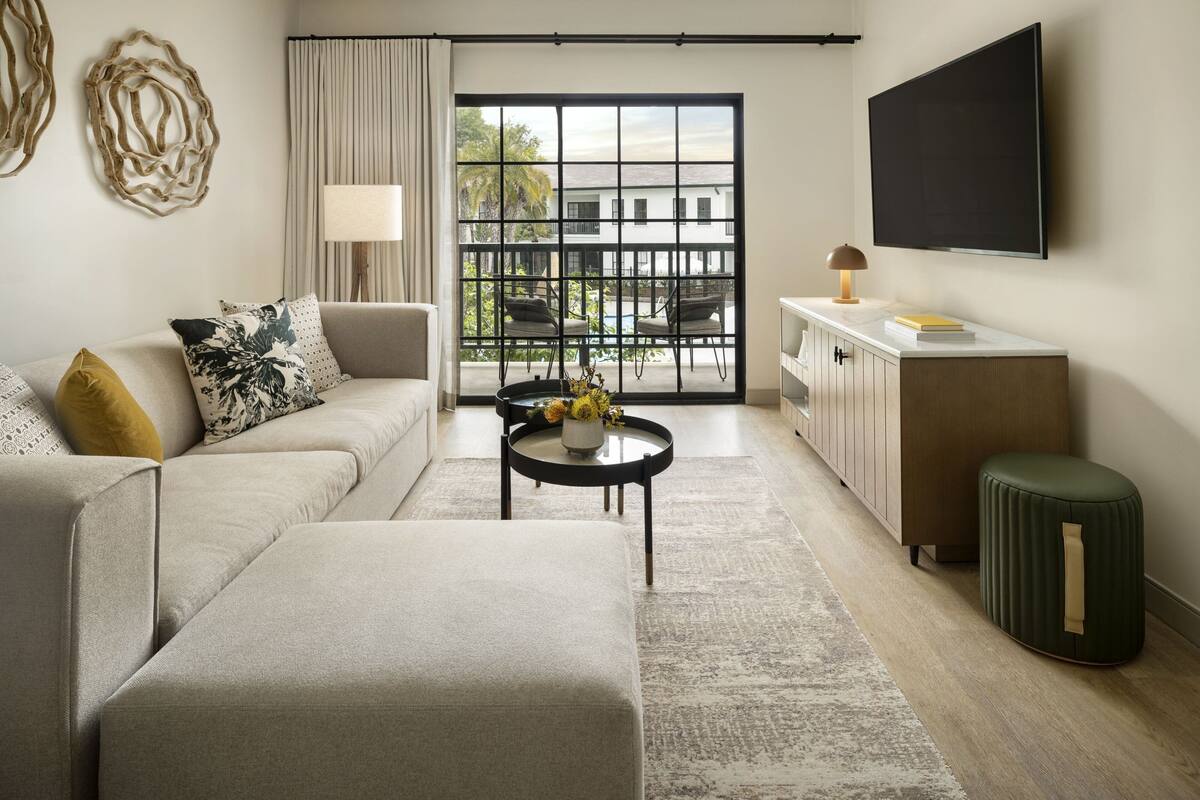 A modern living room with a sofa, ottoman, coffee table, and a view through a large window to the outdoors.