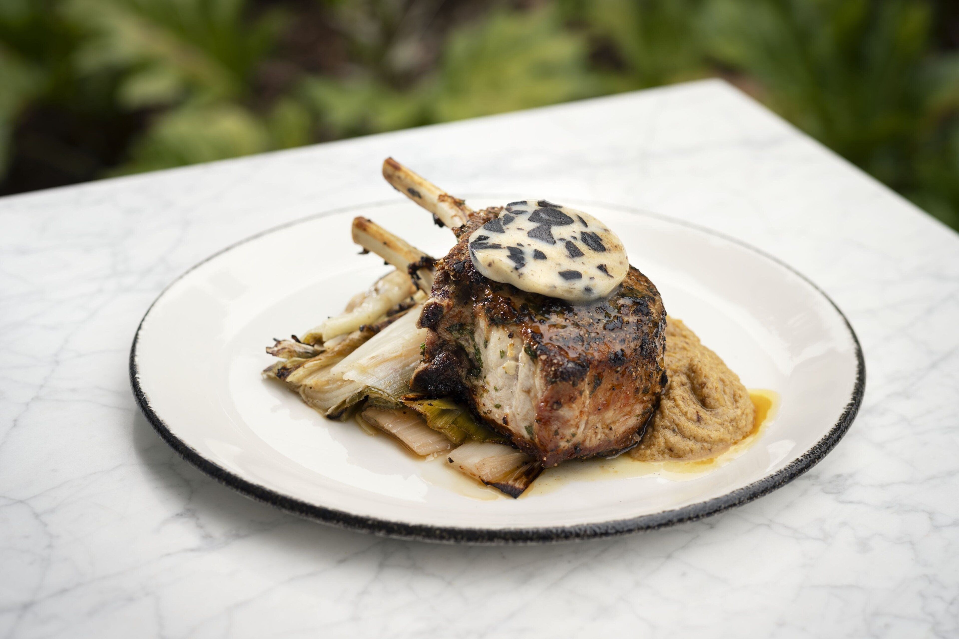 Un plato con un chuletón de cordero, un hongo y algunas verduras sobre una mesa de mármol.