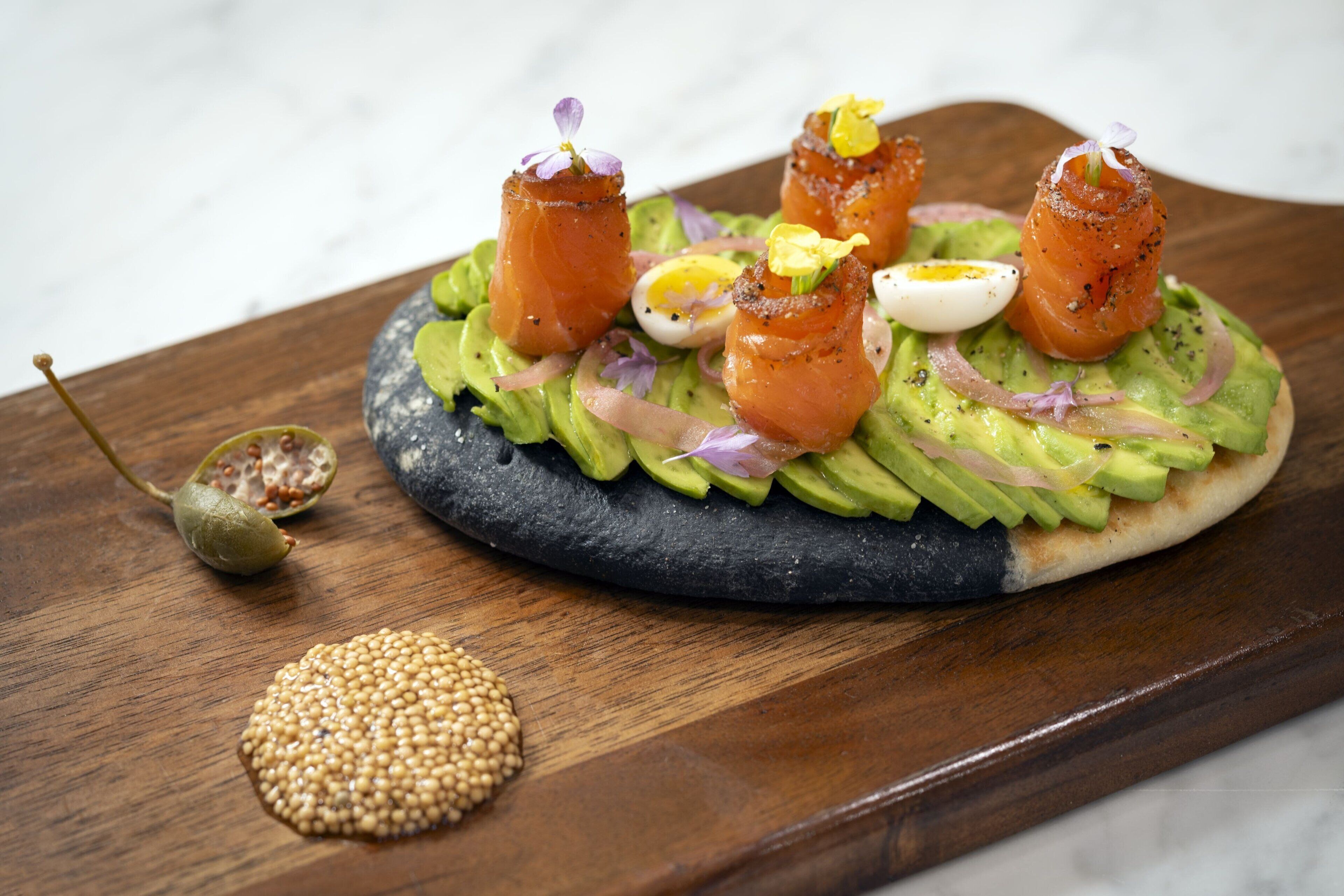 A round flatbread topped with avocado slices, smoked salmon, a boiled egg, and garnished with microgreens and red onion.