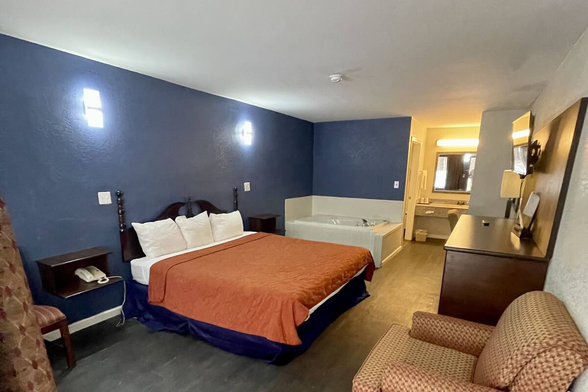 A hotel room with a bed, a chair, a small table, a desk, a mirror, and a bathroom visible through an open door.