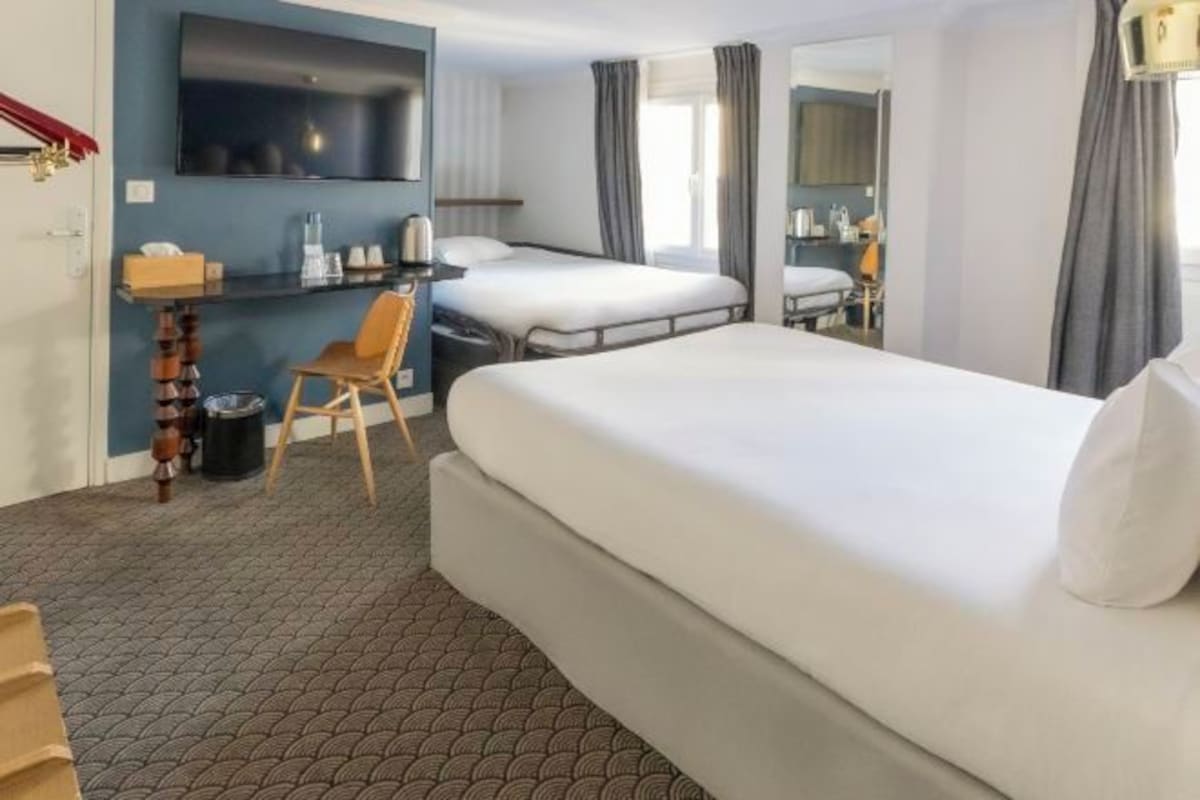 A hotel room with a large bed, a desk with a chair, a flat-screen TV mounted on the wall, and a small trash bin.