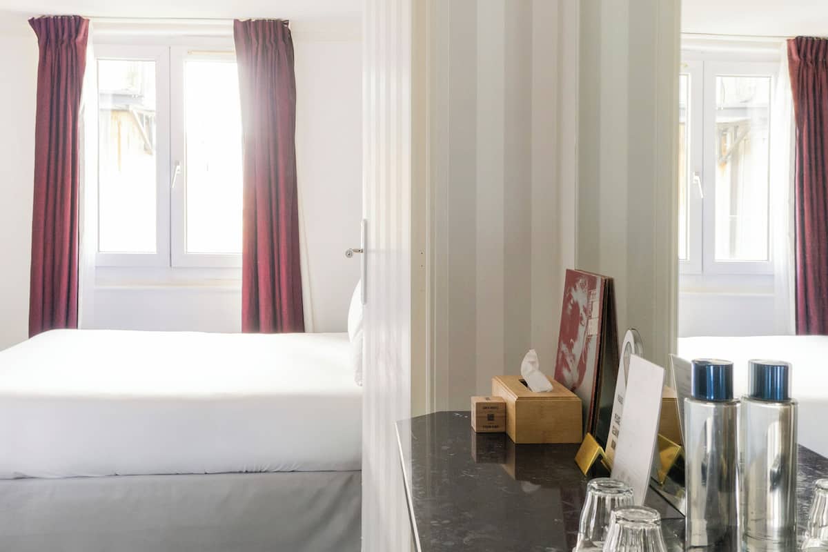 Een hotelkamer met twee bedden, een tafel met marmeren blad en toiletartikelen, en een raam met gordijnen.