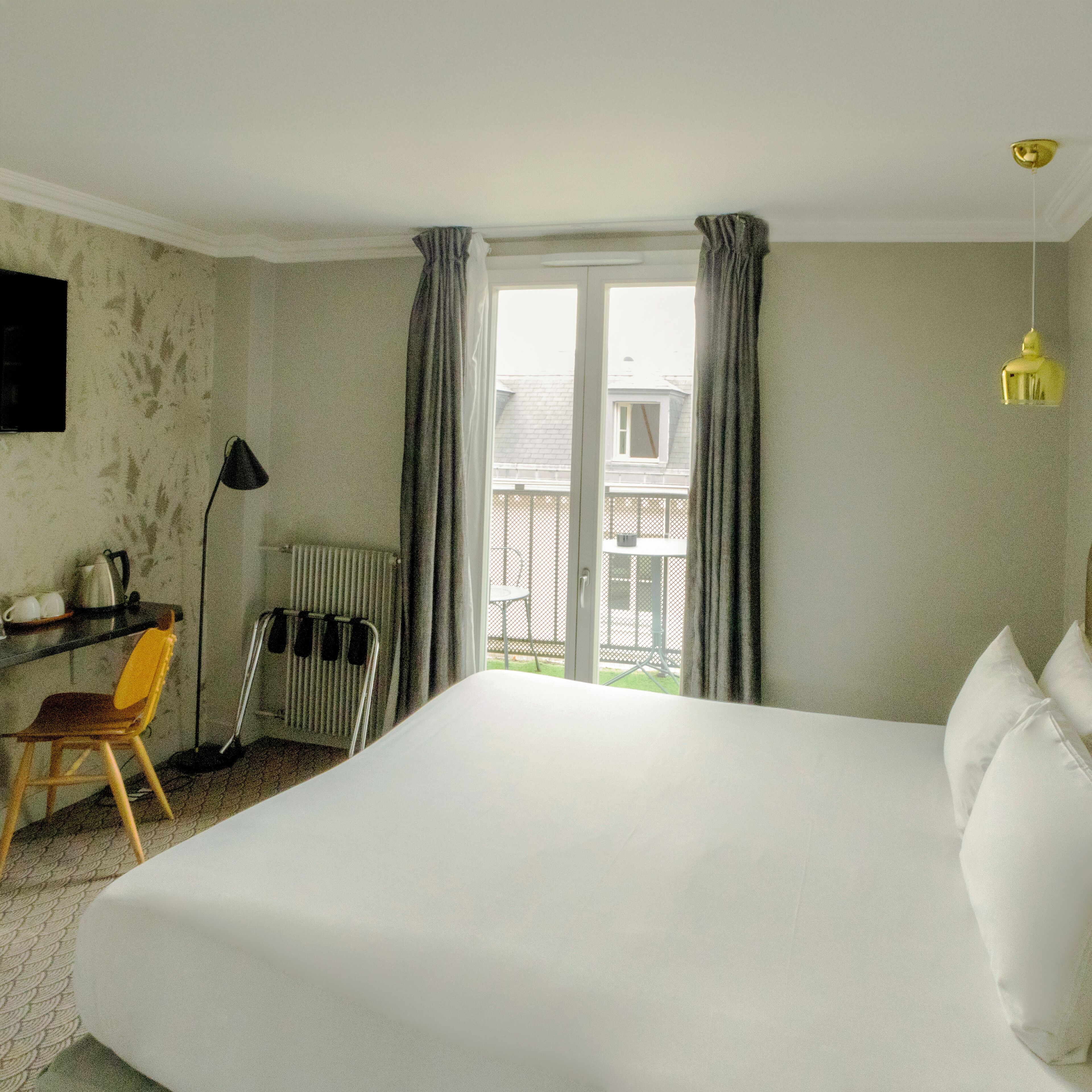 A hotel room with a large bed, a desk with a chair, a radiator, and a window with curtains.