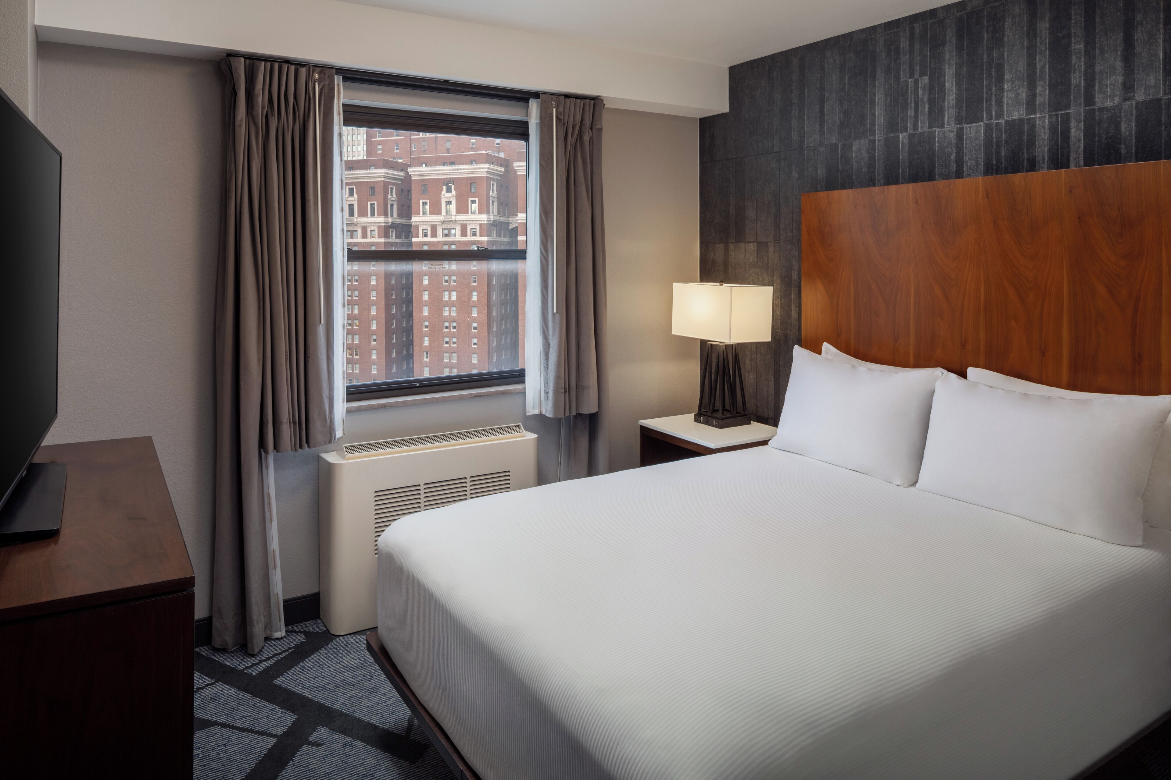 Habitación de hotel con una cama grande, un televisor sobre un escritorio de madera, una ventana con cortinas, una lámpara y vistas a edificios.