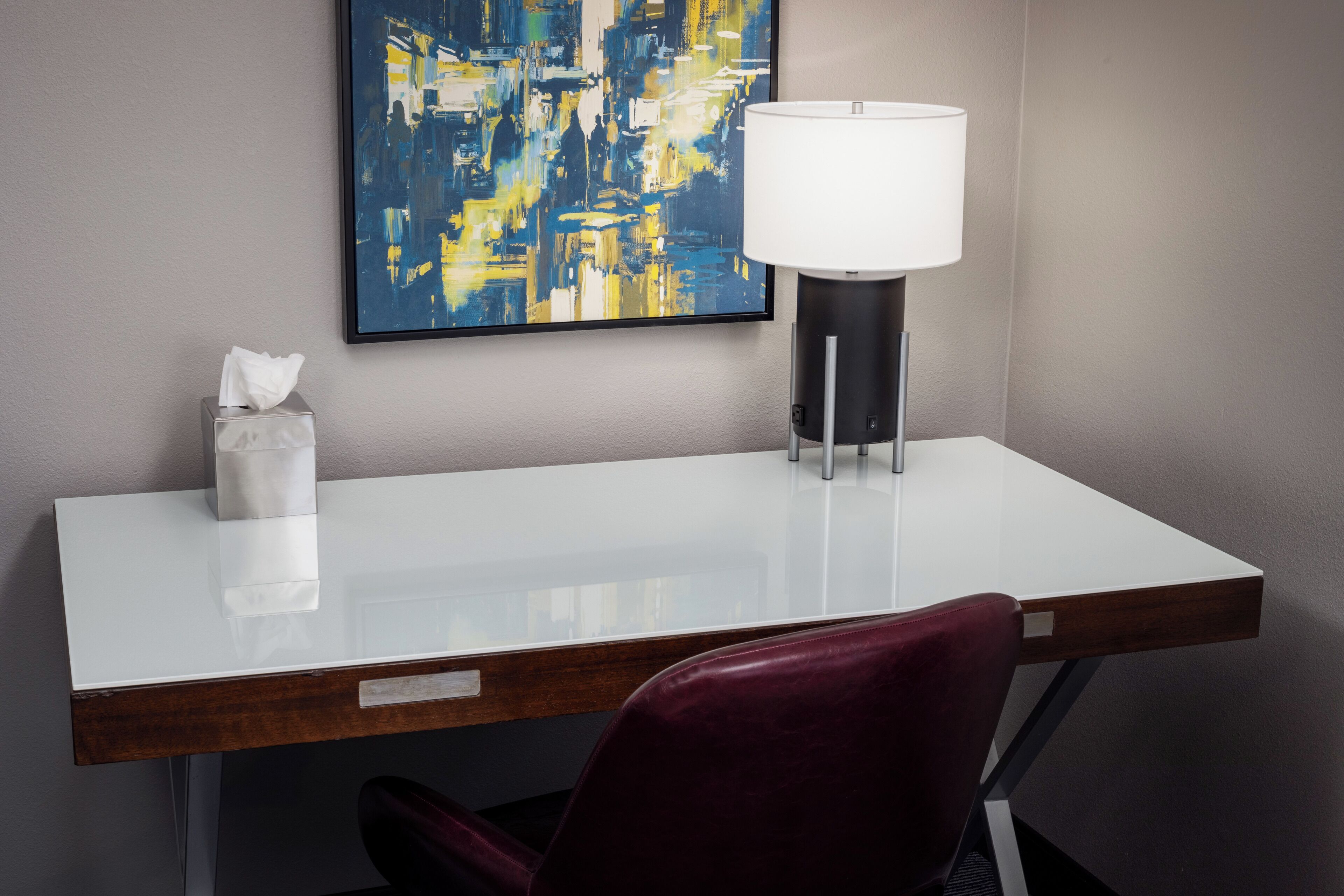A hotel room with a white desk, a red chair, a wall-mounted abstract painting, and a floor lamp.