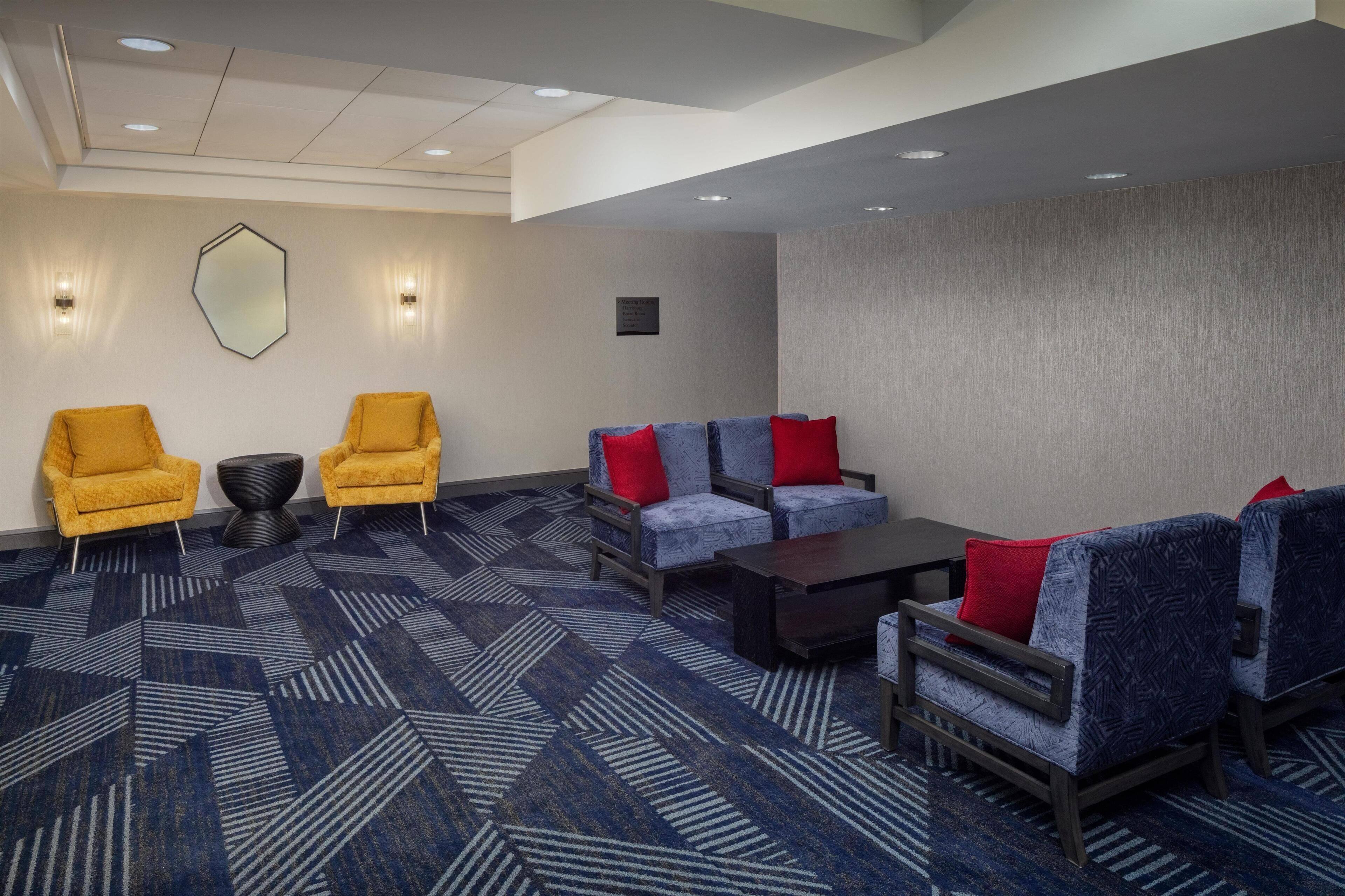 A hotel lobby with a geometric patterned carpet, two yellow armchairs, a blue sofa with red cushions, and a dark coffee table.