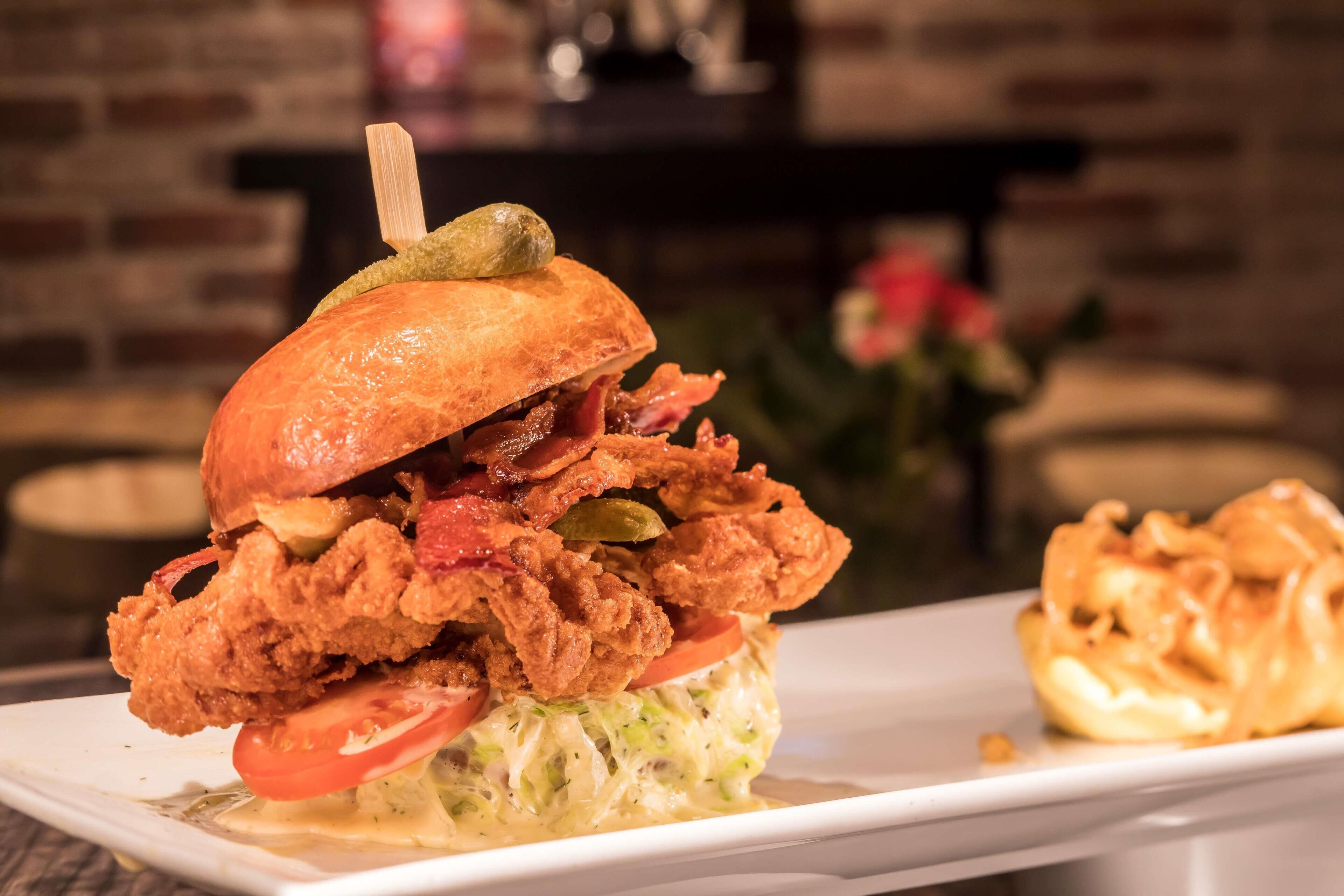 A sandwich with fried chicken, bacon, and a pickle on a bun, served on a white plate with a side of fries.