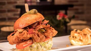 A sandwich with fried chicken, bacon, and a pickle on a bun, served on a white plate with a side of fries.