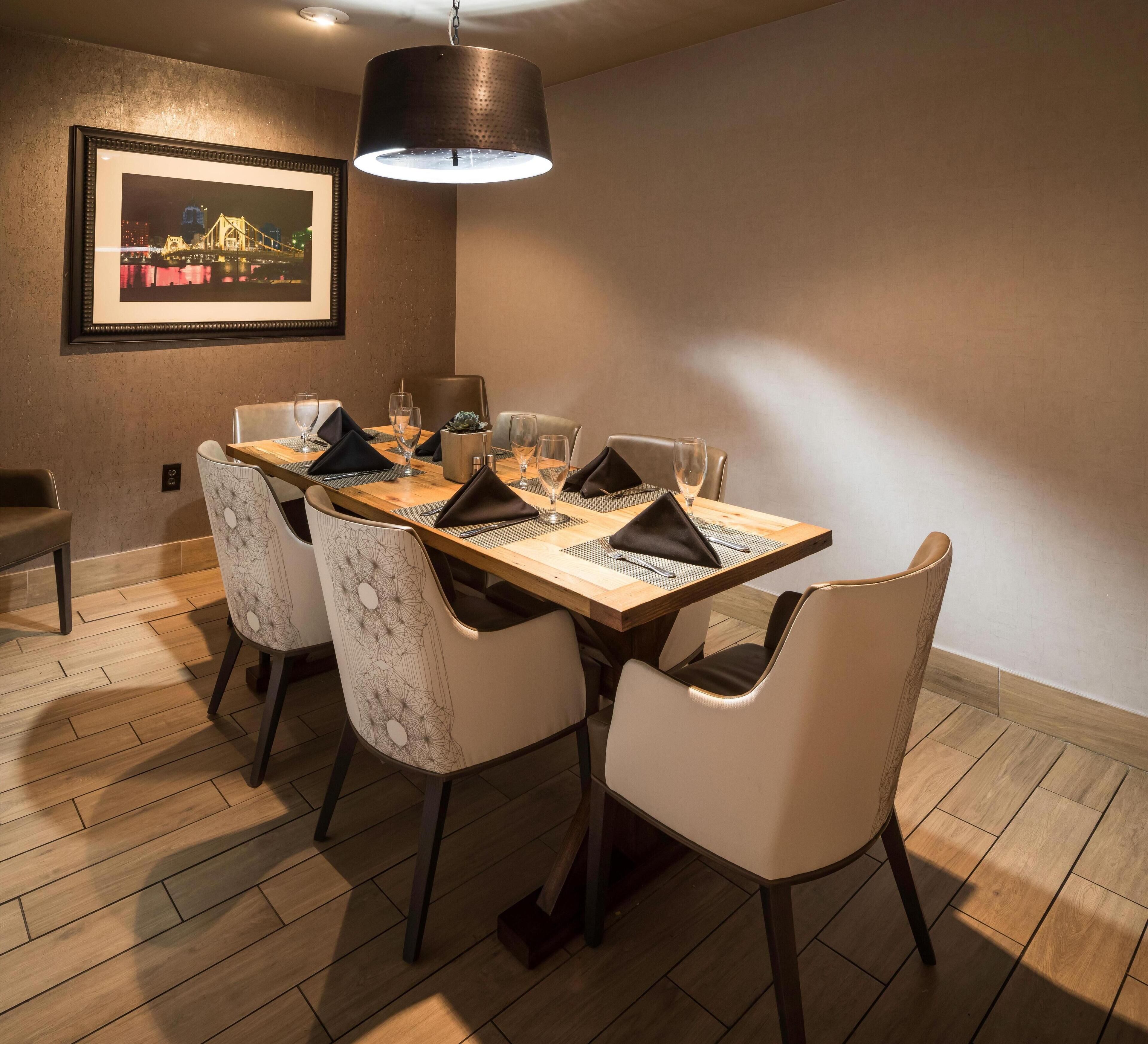 A dining area with a wooden table set for four, surrounded by chairs with geometric patterns. A framed picture of a bridge is on the wall, and a pendant light hangs above the table.