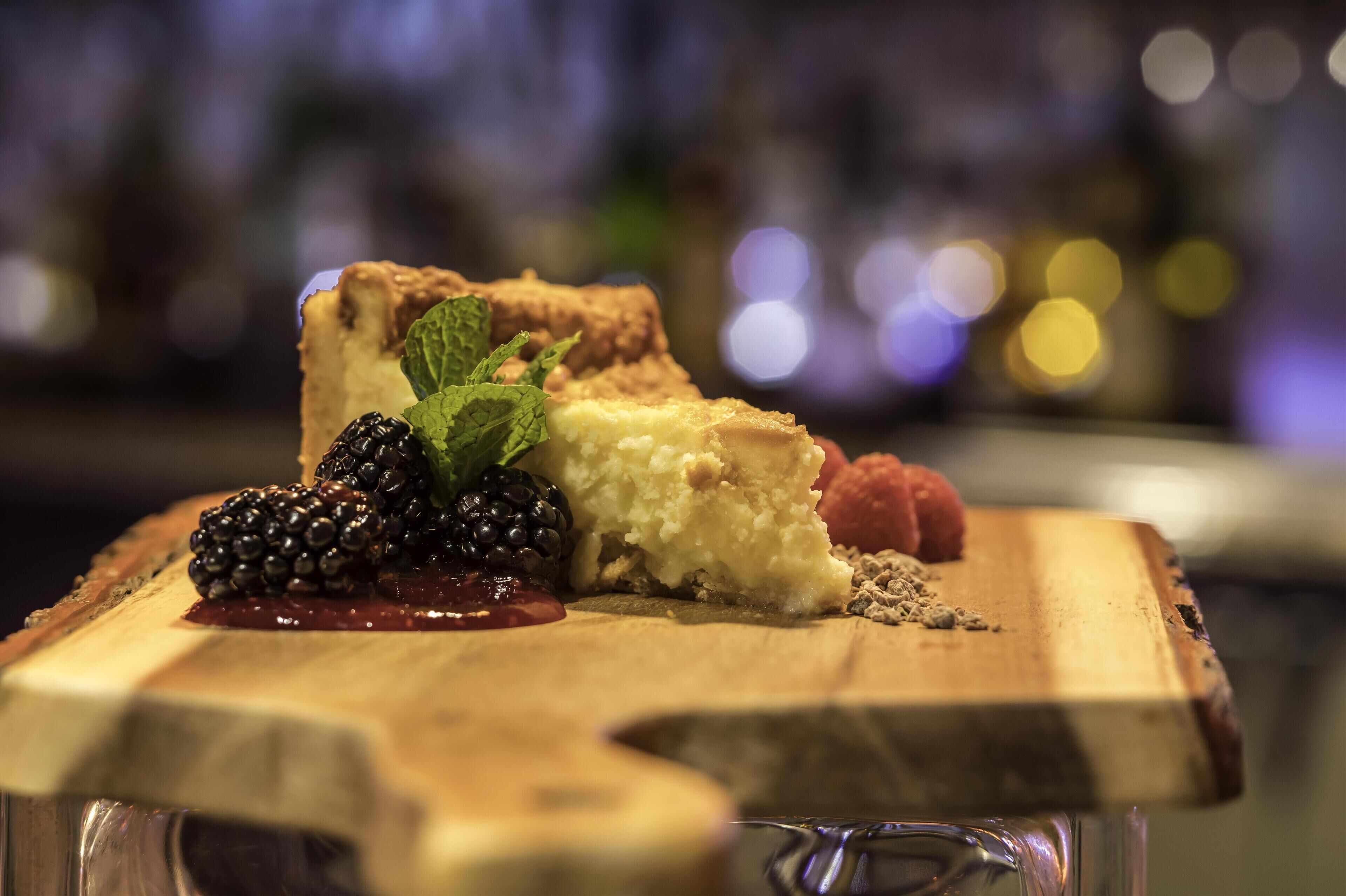A dessert on a wooden board with a slice of cheesecake, blackberries, raspberries, and mint leaves.