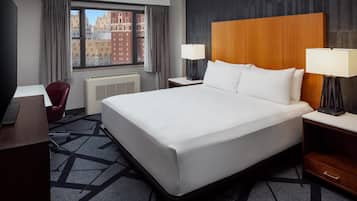 A hotel room with a large bed, two bedside tables with lamps, a desk, a chair, and a window with a view of buildings.