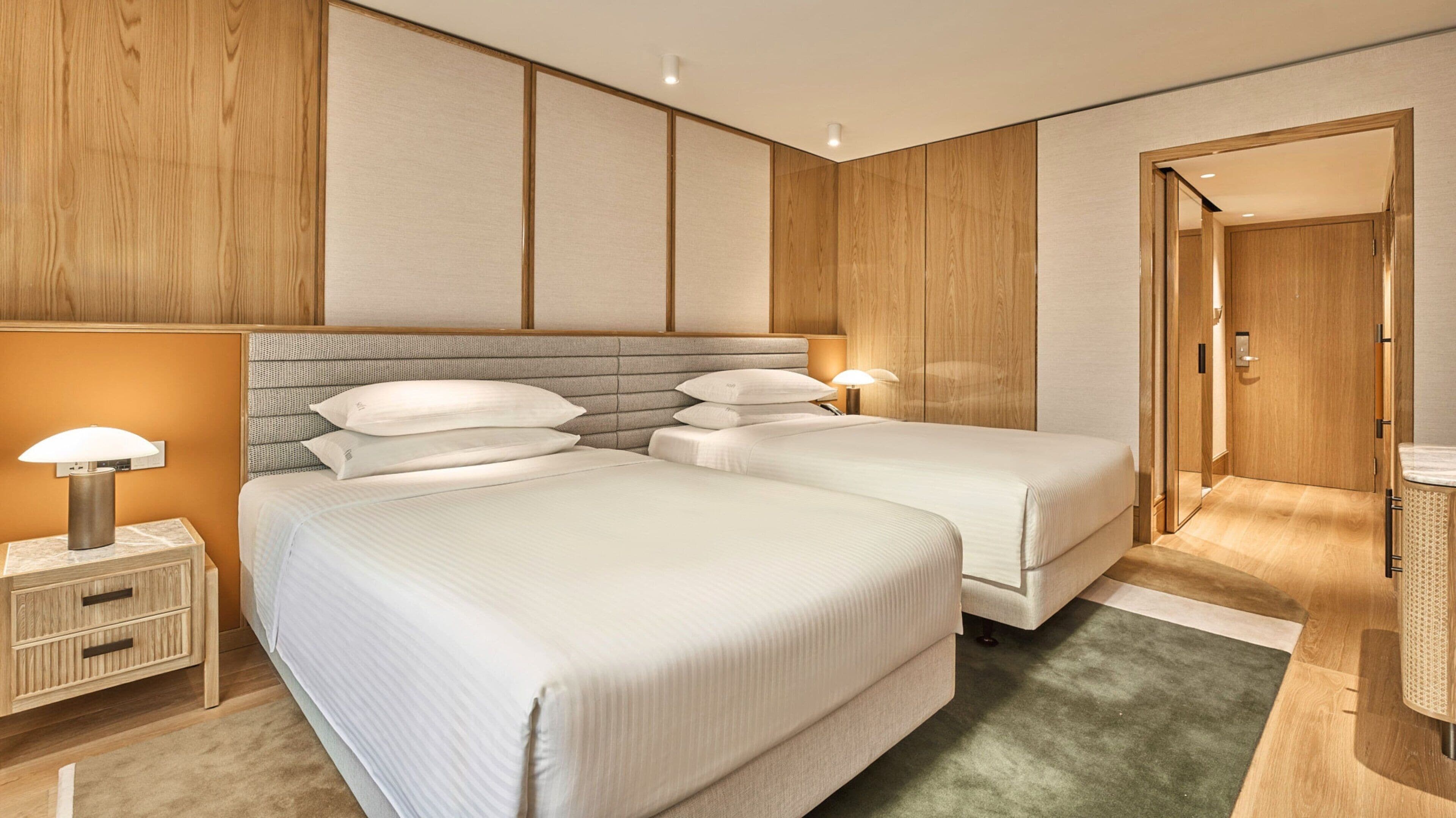 Une chambre d’hôtel avec deux lits, une table de chevet avec une lampe, une tête de lit en bois et une porte donnant sur une autre pièce.