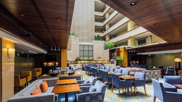 A spacious hotel lobby with multiple seating areas, a central staircase, and a large TV screen.