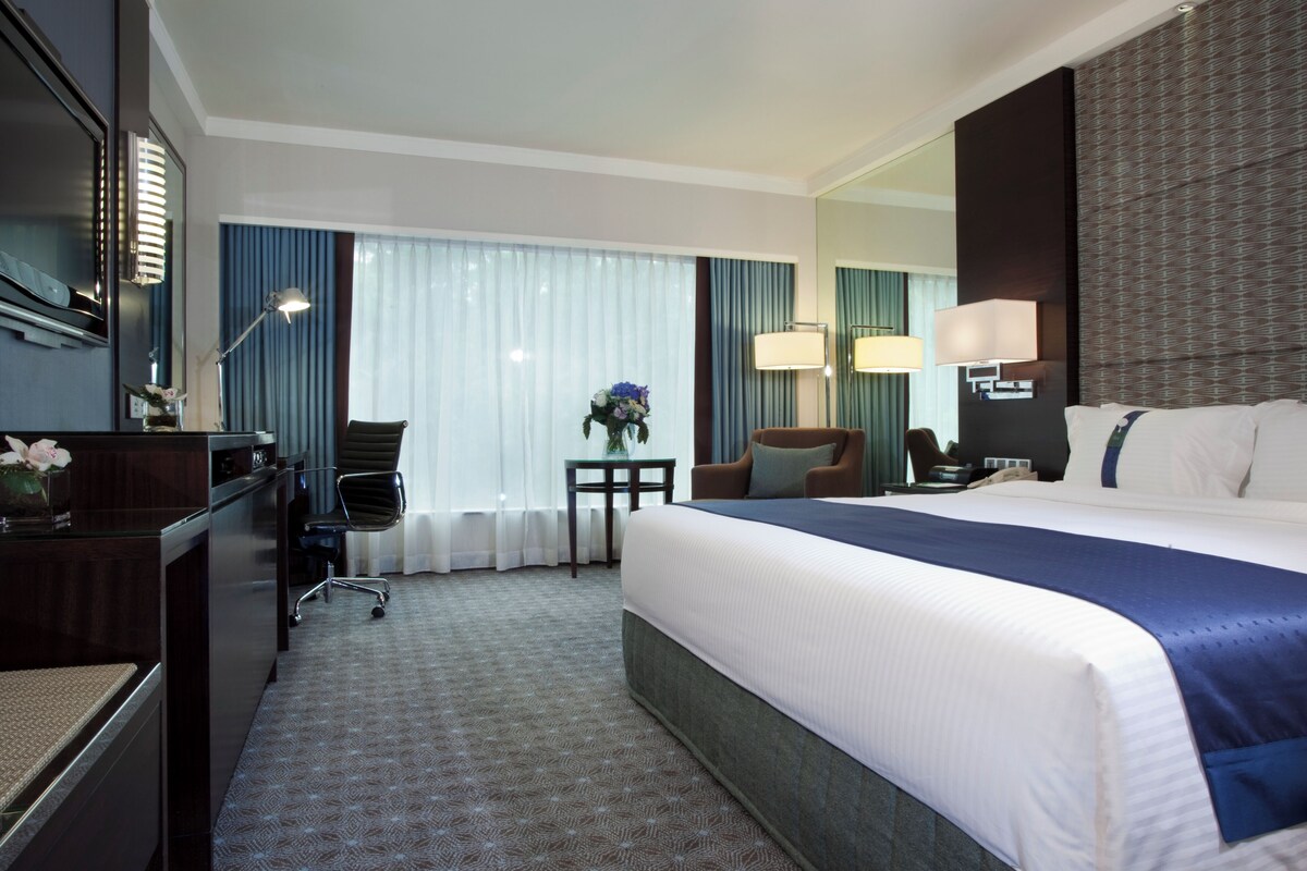 Habitación de hotel con una cama grande, un escritorio, una silla, una mesita con un florero y un espejo en la pared.