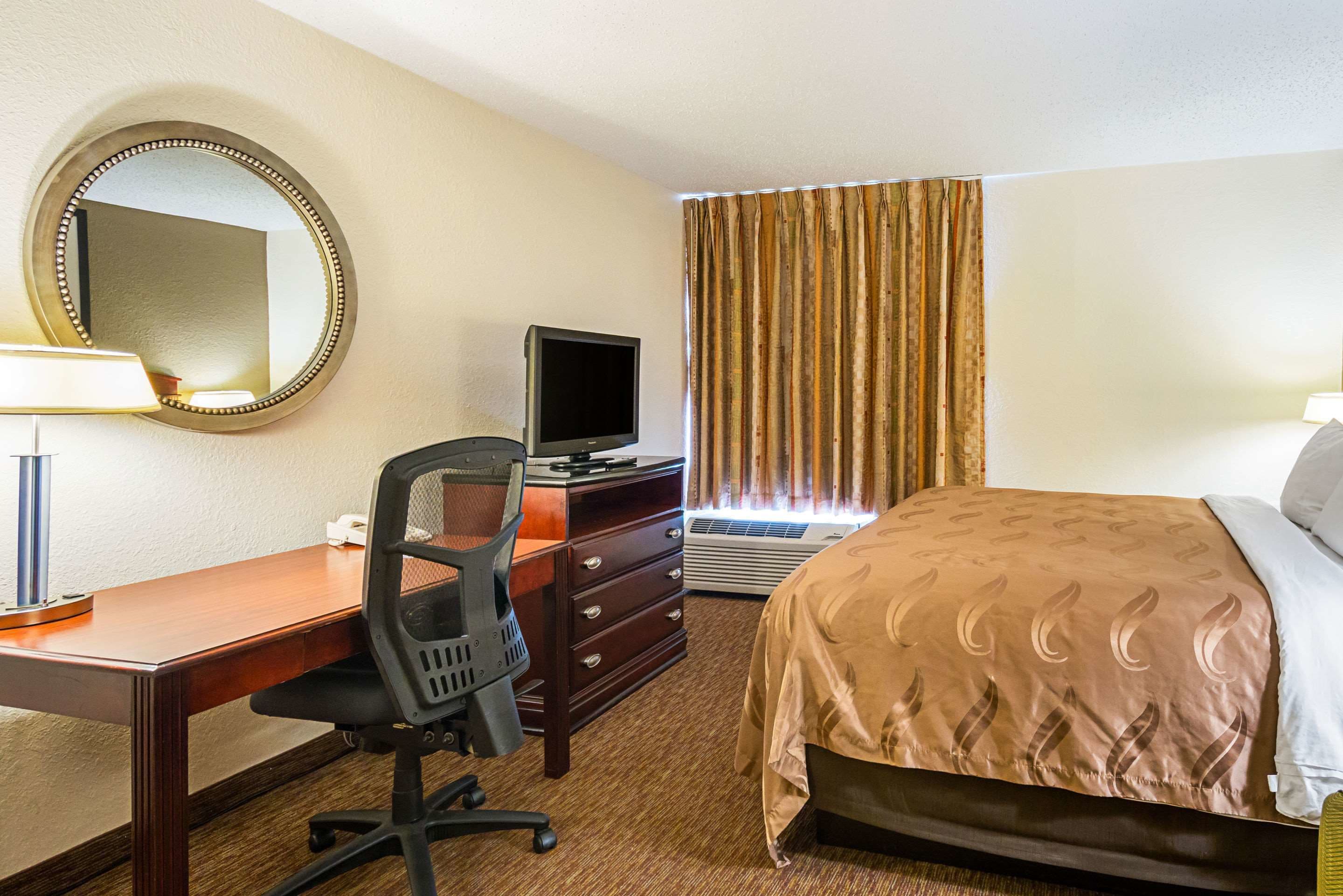 Una habitación de hotel con cama, escritorio con silla, televisión y ventana con cortinas.