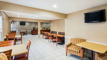 A hotel dining area with multiple tables and chairs, a television mounted on the wall, and a kitchen in the background.