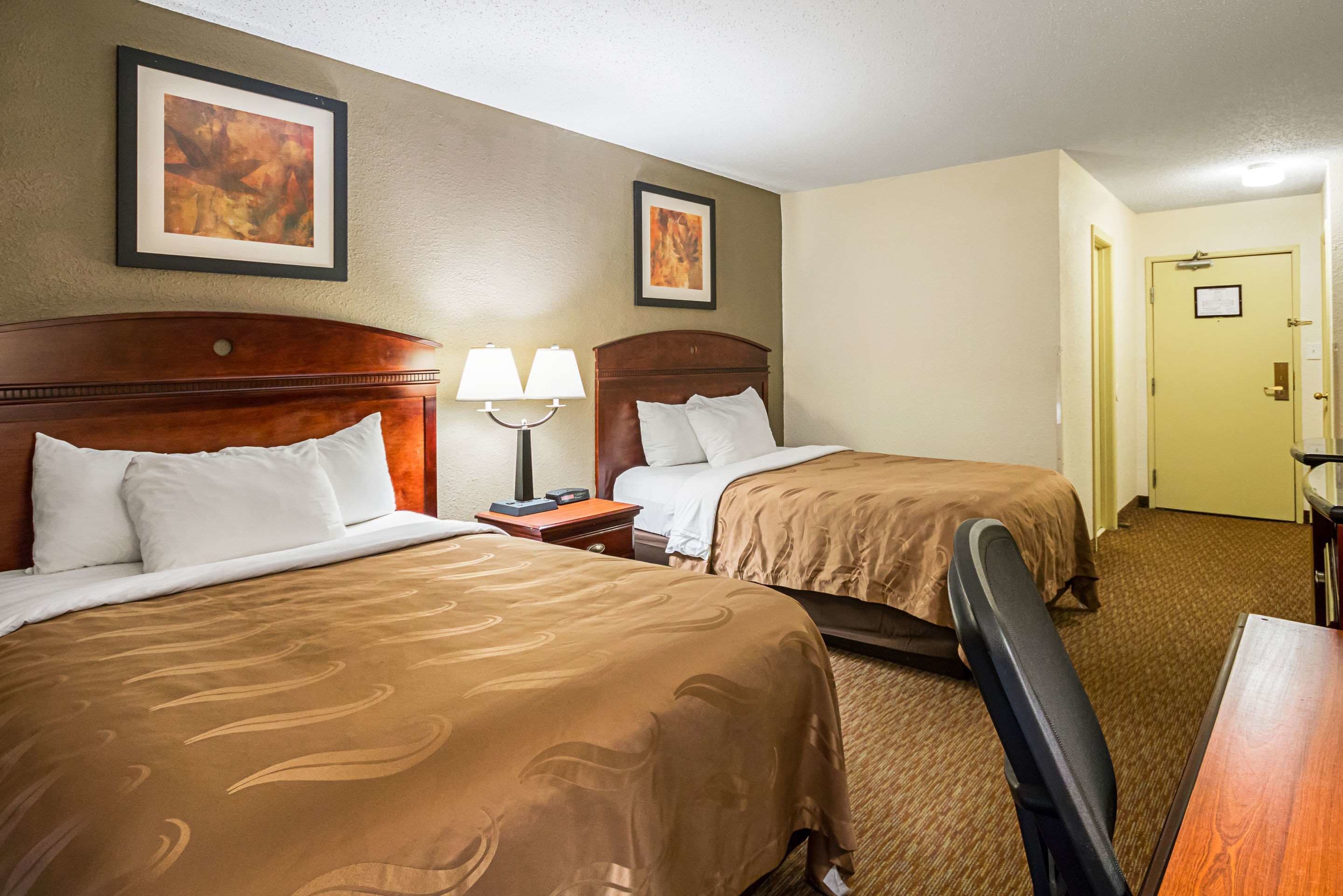 Quarto de hotel com duas camas, uma escrivaninha, uma cadeira e duas fotos emolduradas na parede.