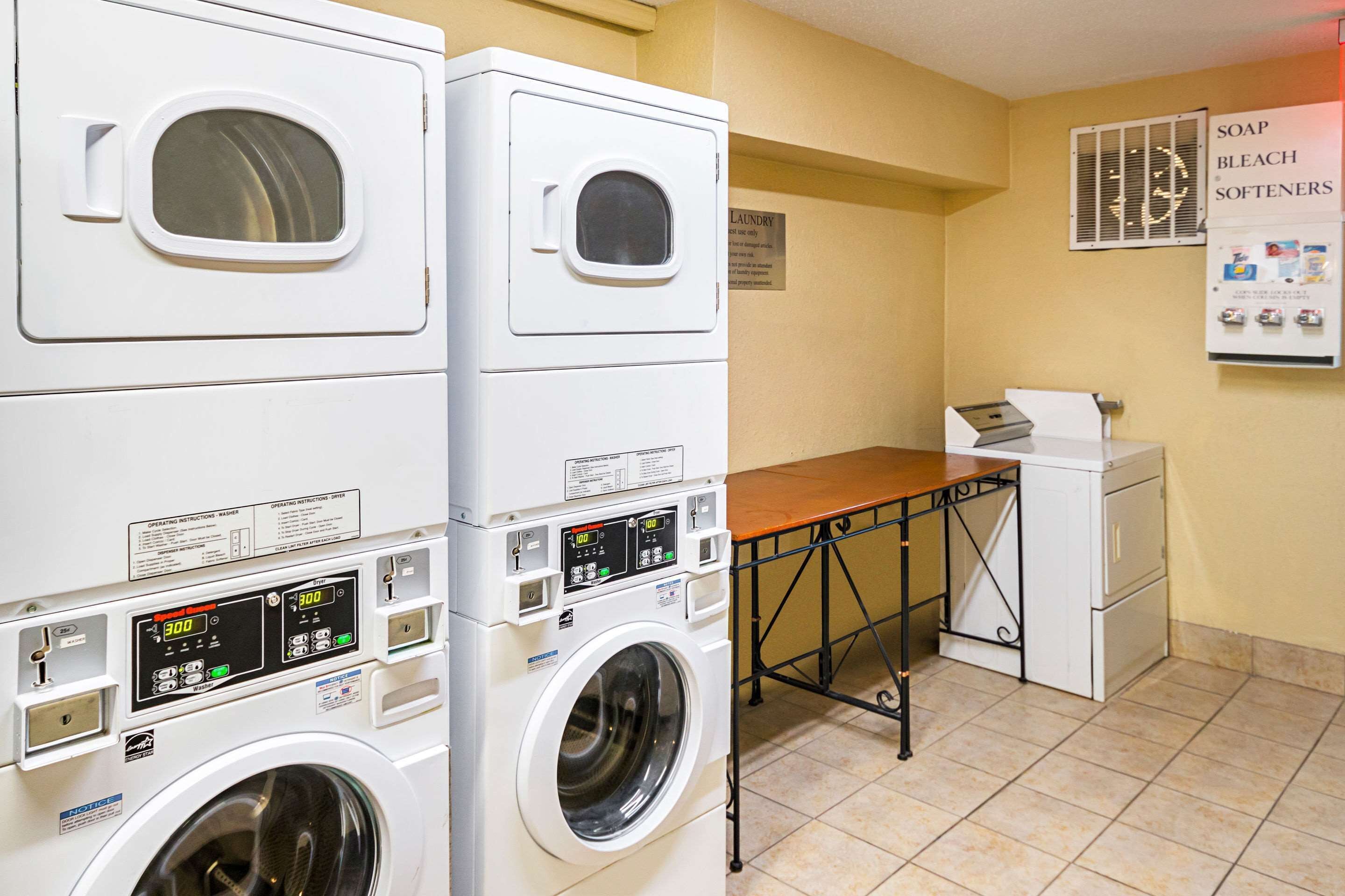 A laundry area with two stacked washers and a dryer, a wooden table, and a sign about soap and bleach softeners.