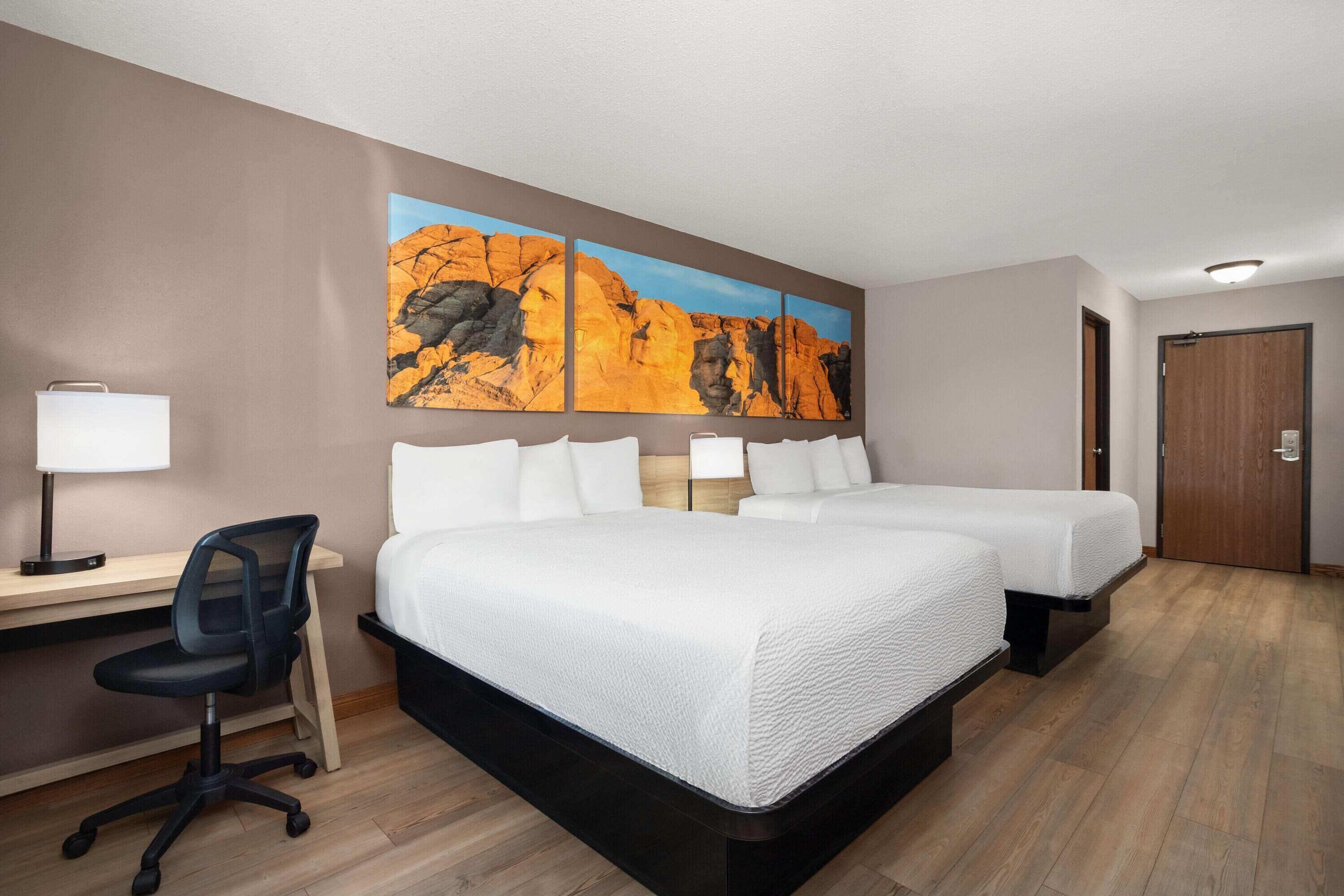 Habitación de hotel con escritorio, dos camas, una silla y un cuadro colgado en la pared.