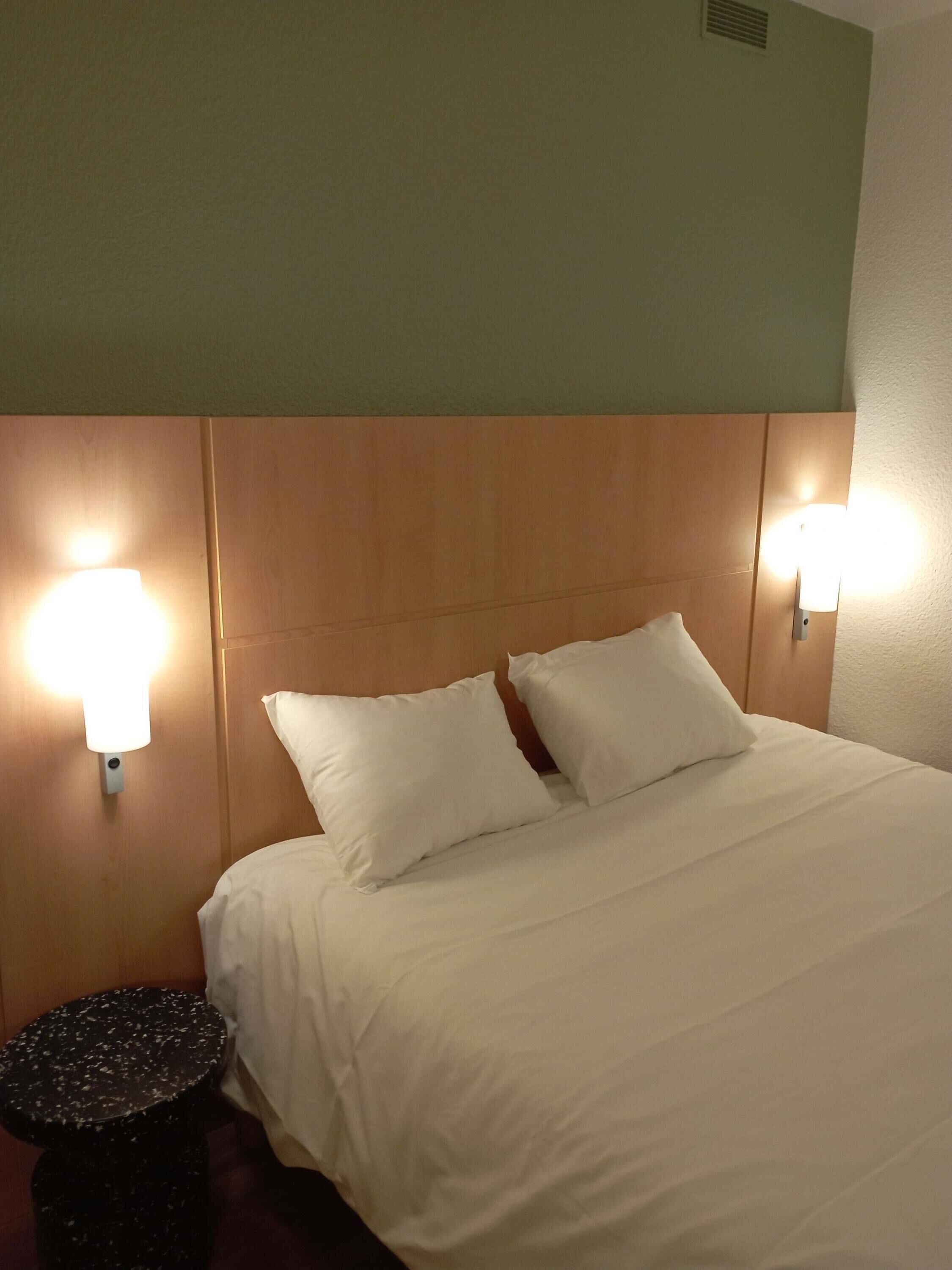 A hotel room with a bed, two wall-mounted lamps, and a round black stool.