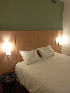 A hotel room with a bed, two bedside lamps, a round black stool, and a wooden headboard. - Ibis Fourmies (Fourmies)