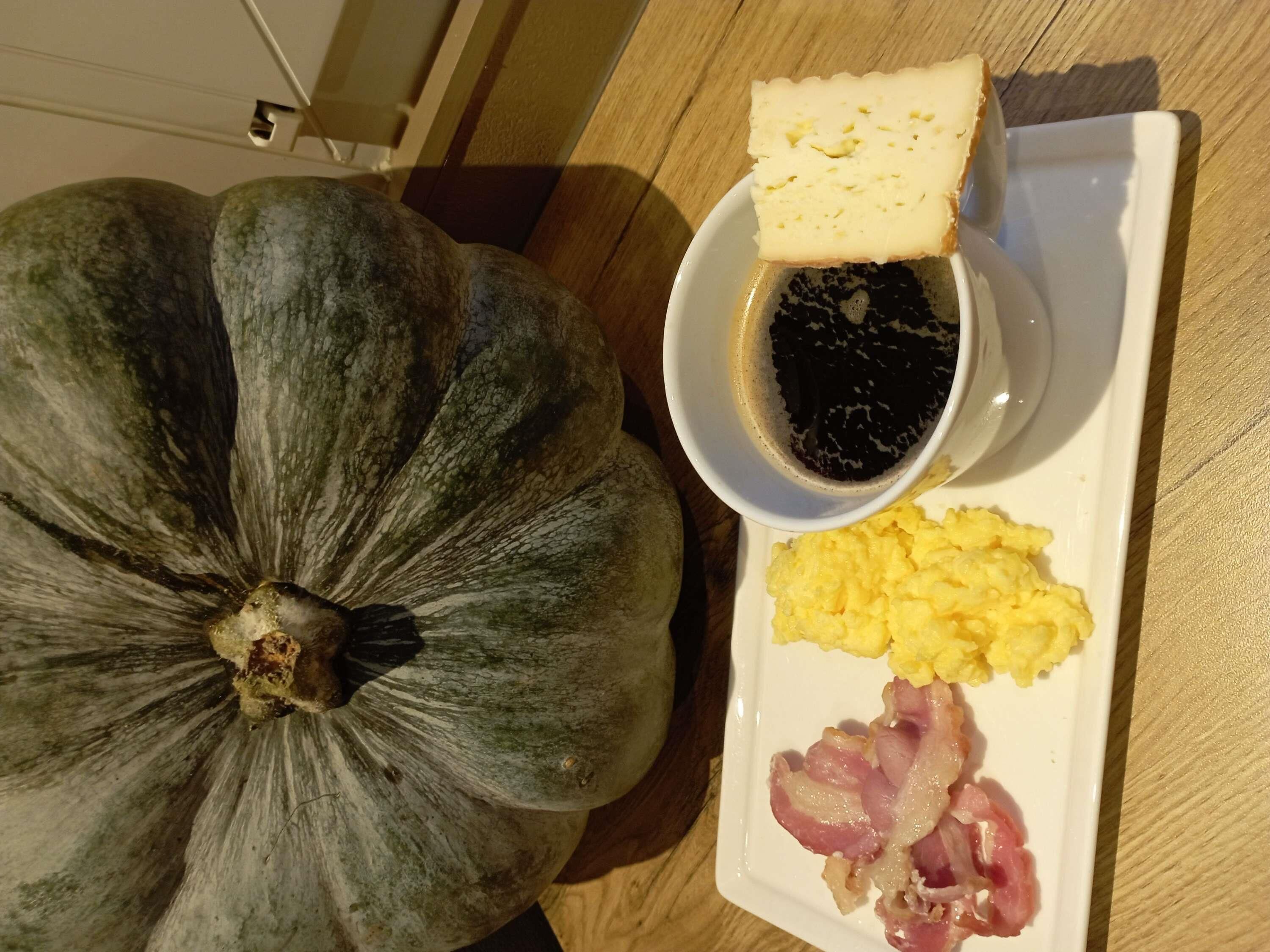Ein Teller mit Rührei, Speck und einer Käsescheibe, dazu eine Schüssel schwarze Soße und ein Kürbis.